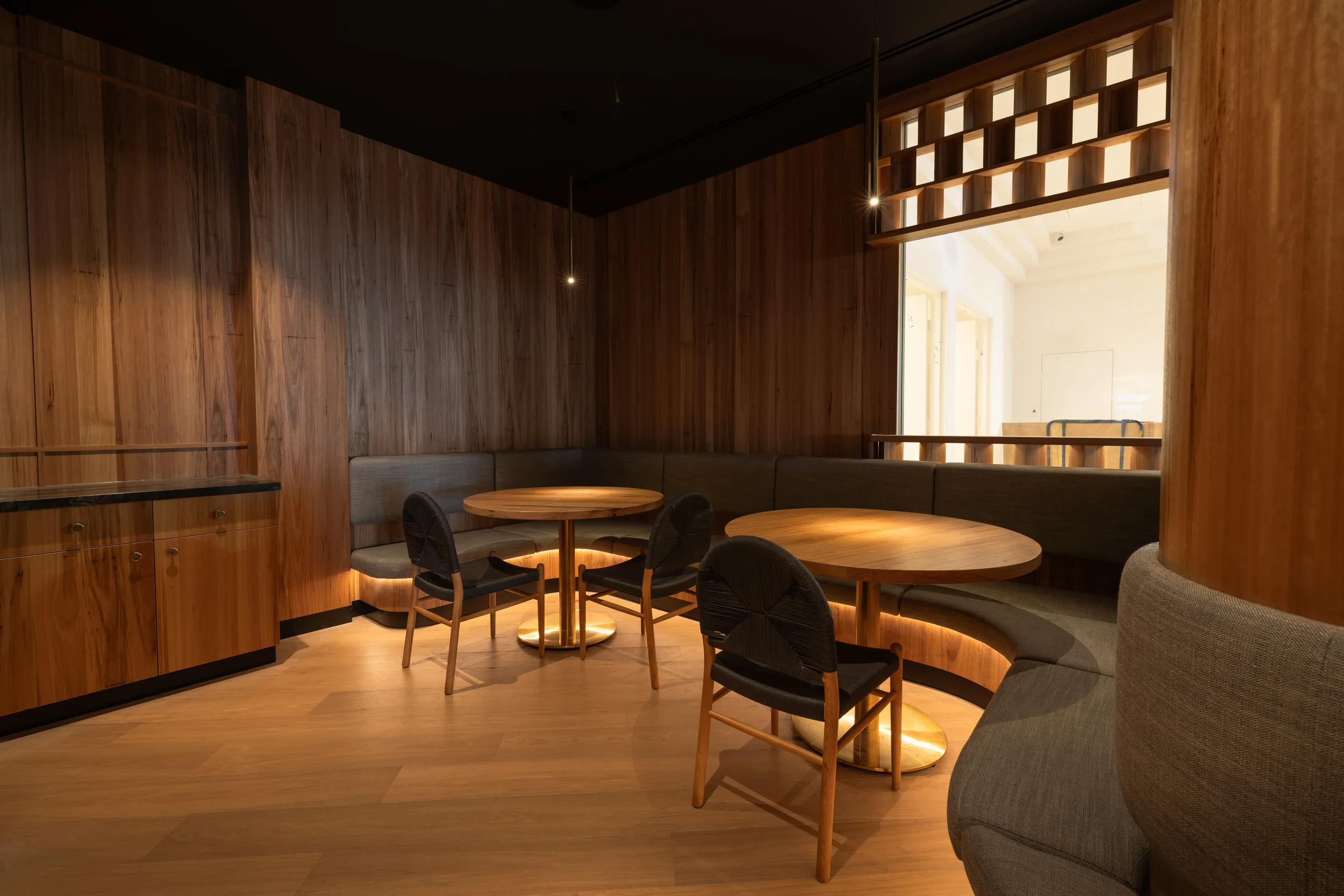 Interior of a room with wood-paneled walls, round wooden tables, and black chairs with rattan backs. There is a cushioned curved seating area along the wall, overhead pendant lighting, and a large window letting in natural light.