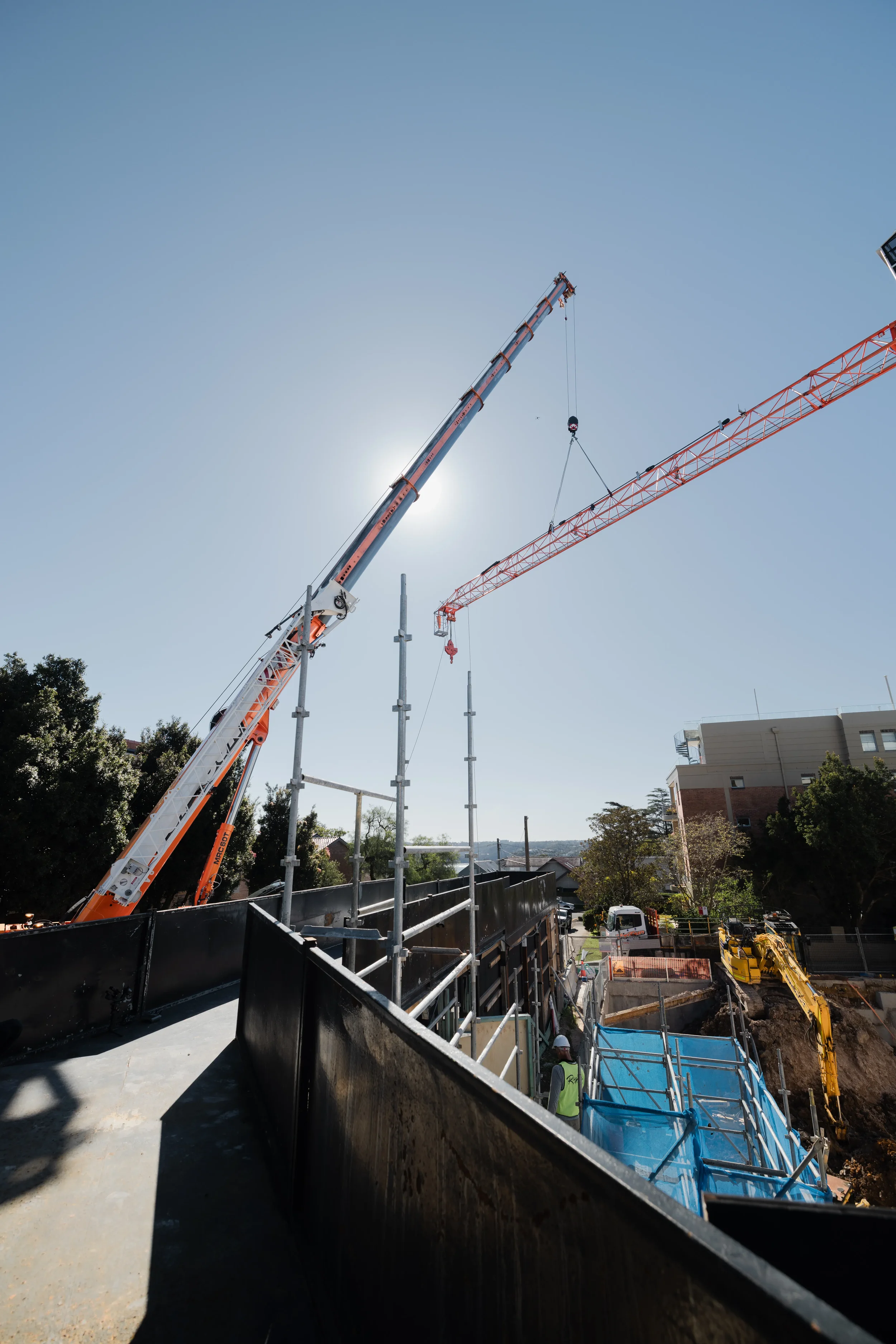 Construction site with cranes, scaffolding, and workers, with trees and buildings in the background.