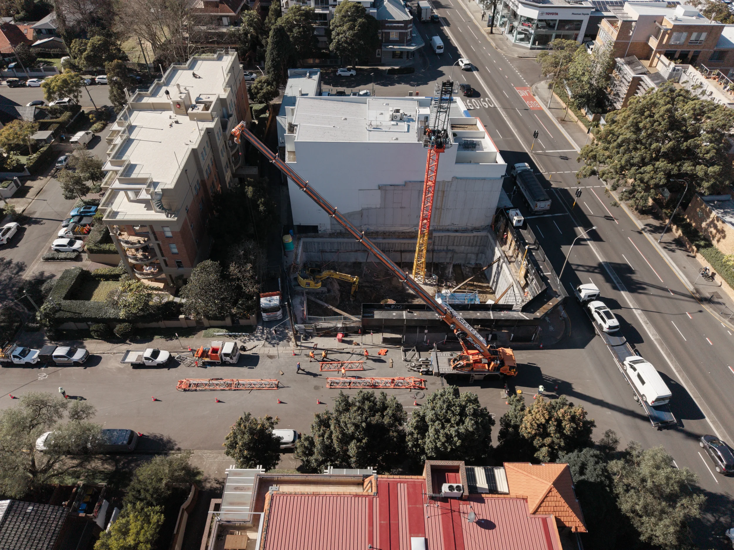 An aerial view of a city street under construction with a crane lifting materials to the top of a building, surrounded by parked cars, trees, and residential and commercial buildings.