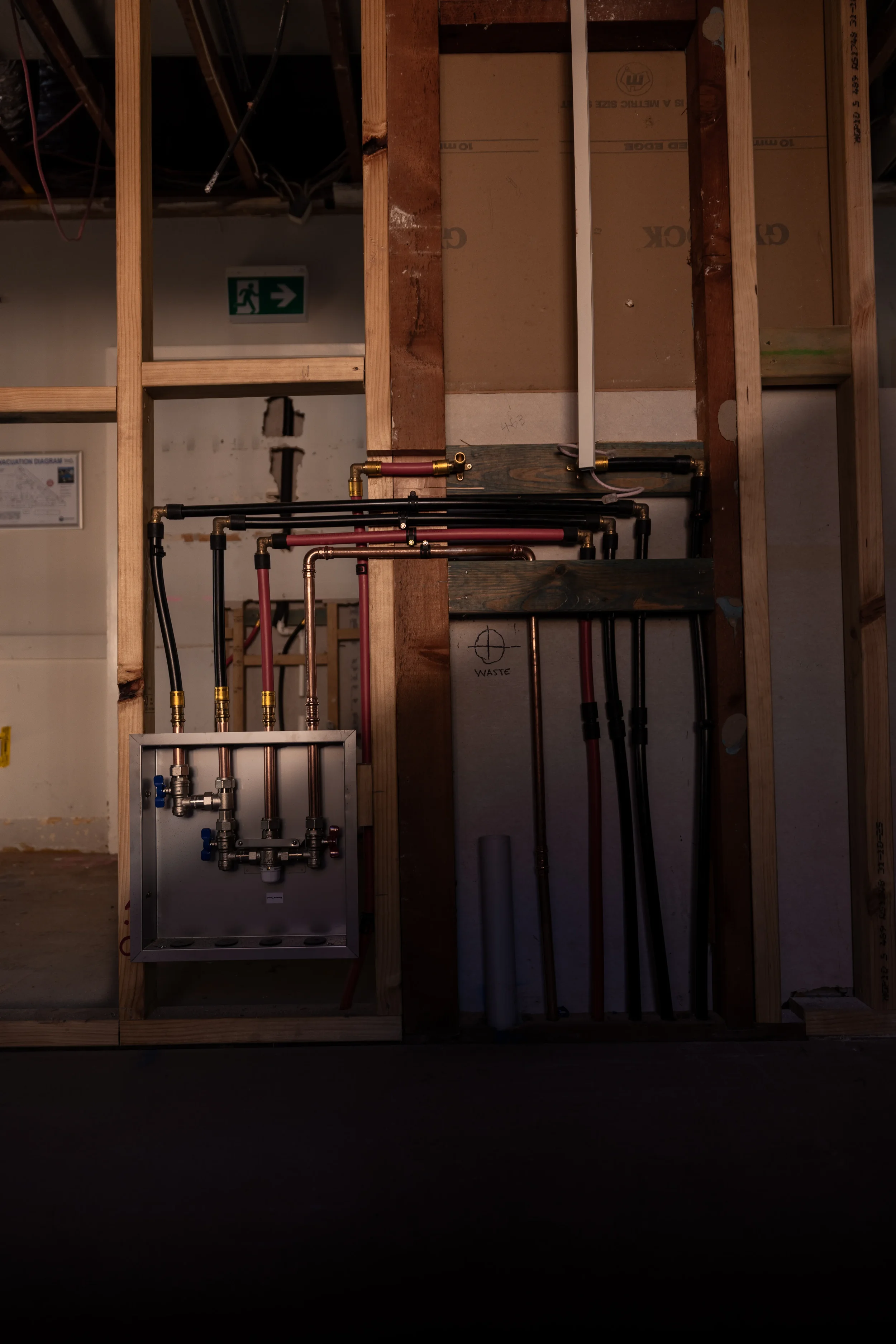 Construction site with exposed wooden framing and plumbing pipes, electrical wiring, and an emergency exit sign in the background.