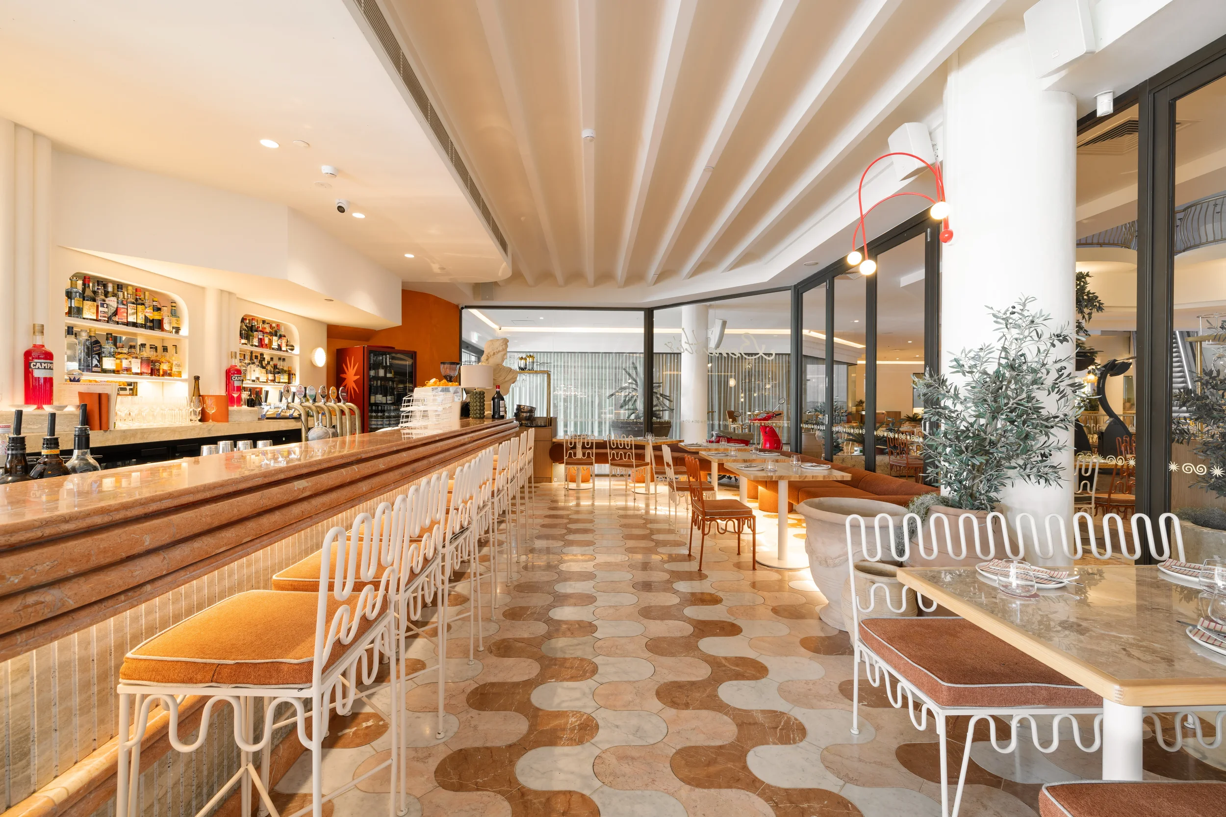 Interior of a stylish, modern restaurant or bar with a marble counter, barstools, tables, and chairs, decorated with plants and contemporary lighting fixtures.