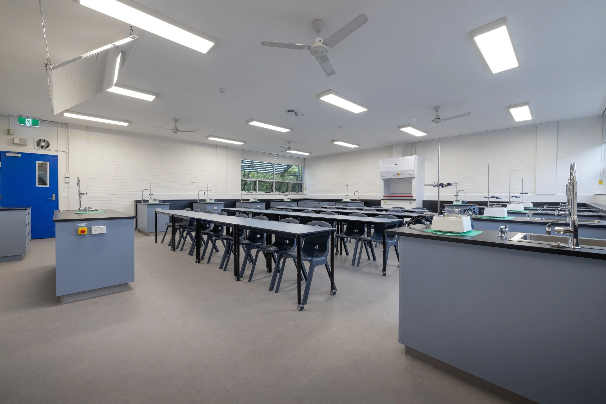 Empty science classroom with gray tables and black chairs, lab sinks, and equipment, bright lighting, white walls, window showing greenery outside.