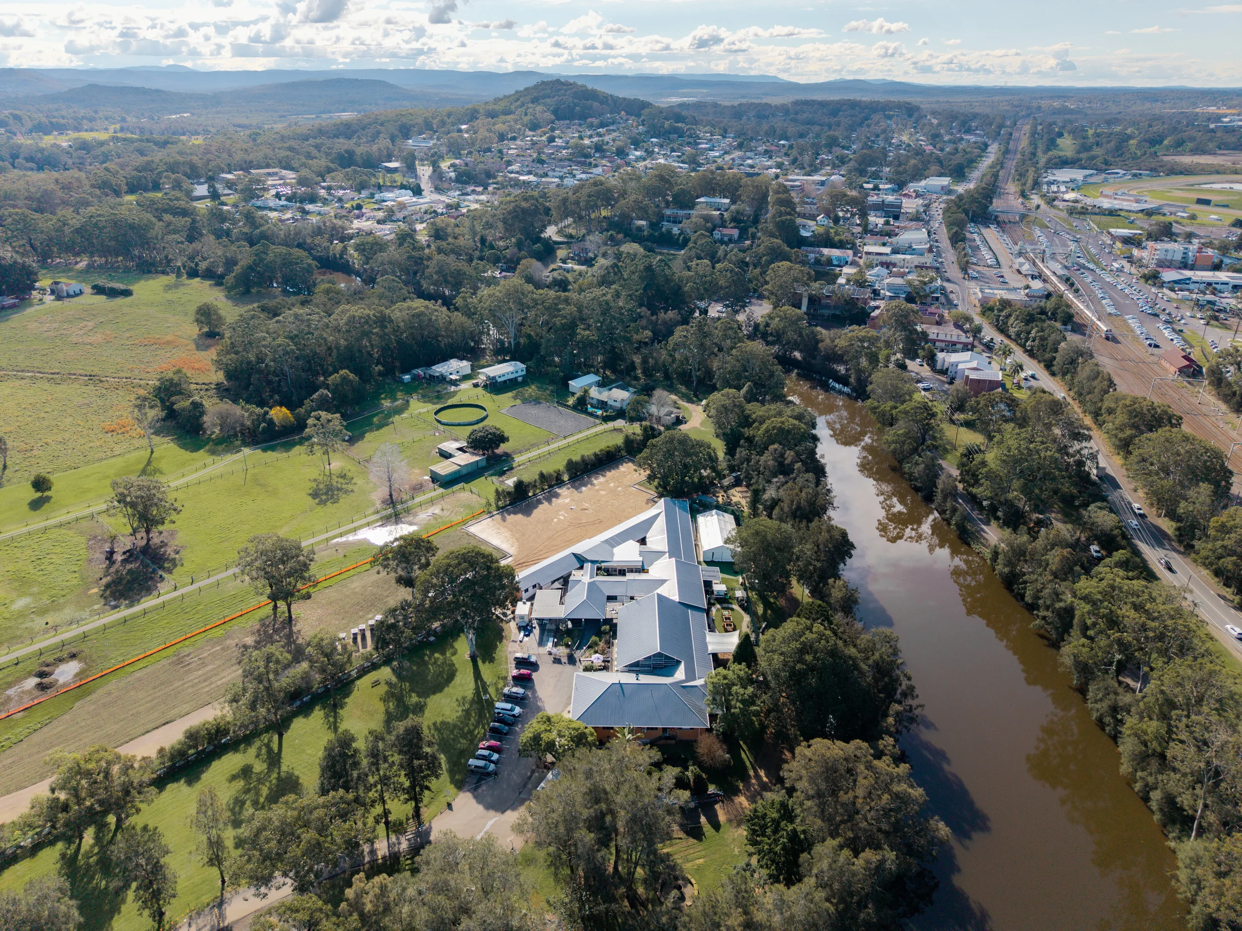 An aerial view of a small town surrounded by greenery, featuring a river running through it, a sports stadium, and residential buildings, under a partly cloudy sky.
