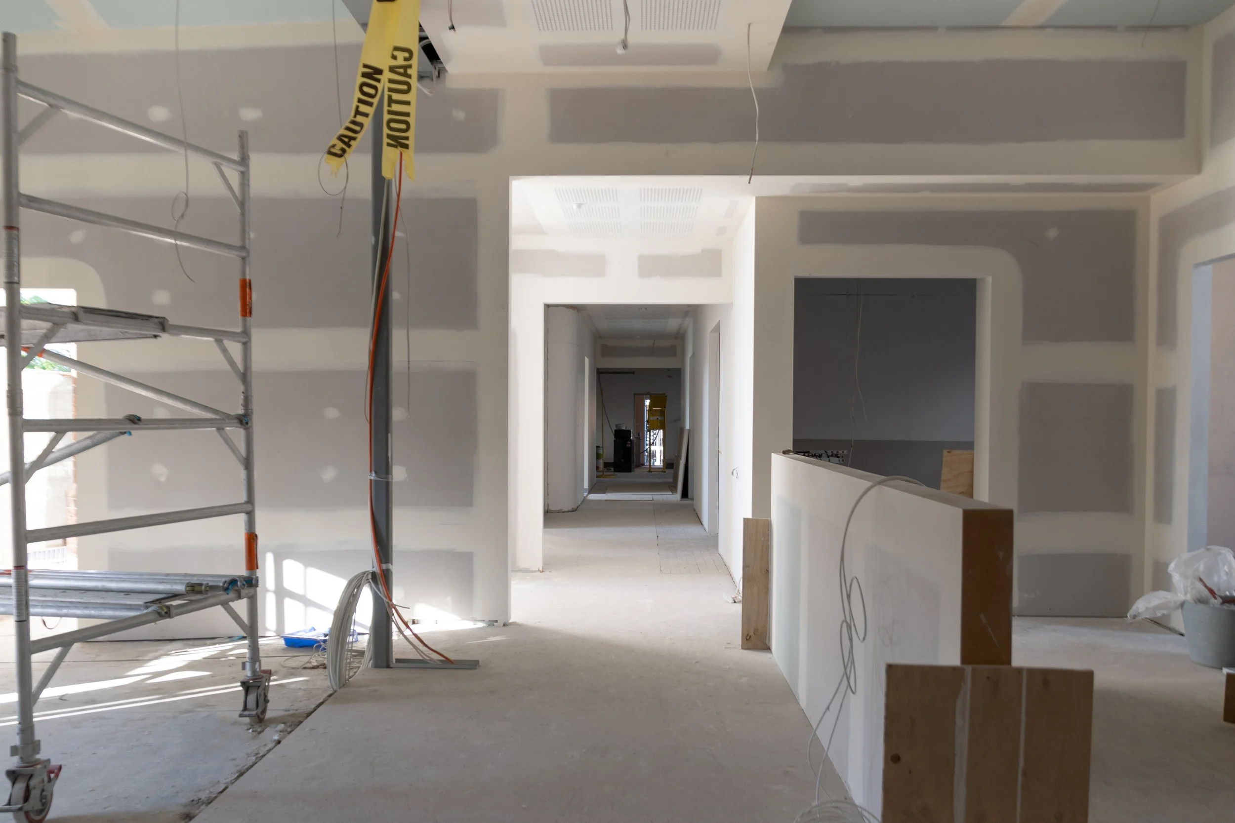 Interior view of a building under construction with drywall walls and ceiling, scaffolding on the left, and electrical wiring hanging from the ceiling.