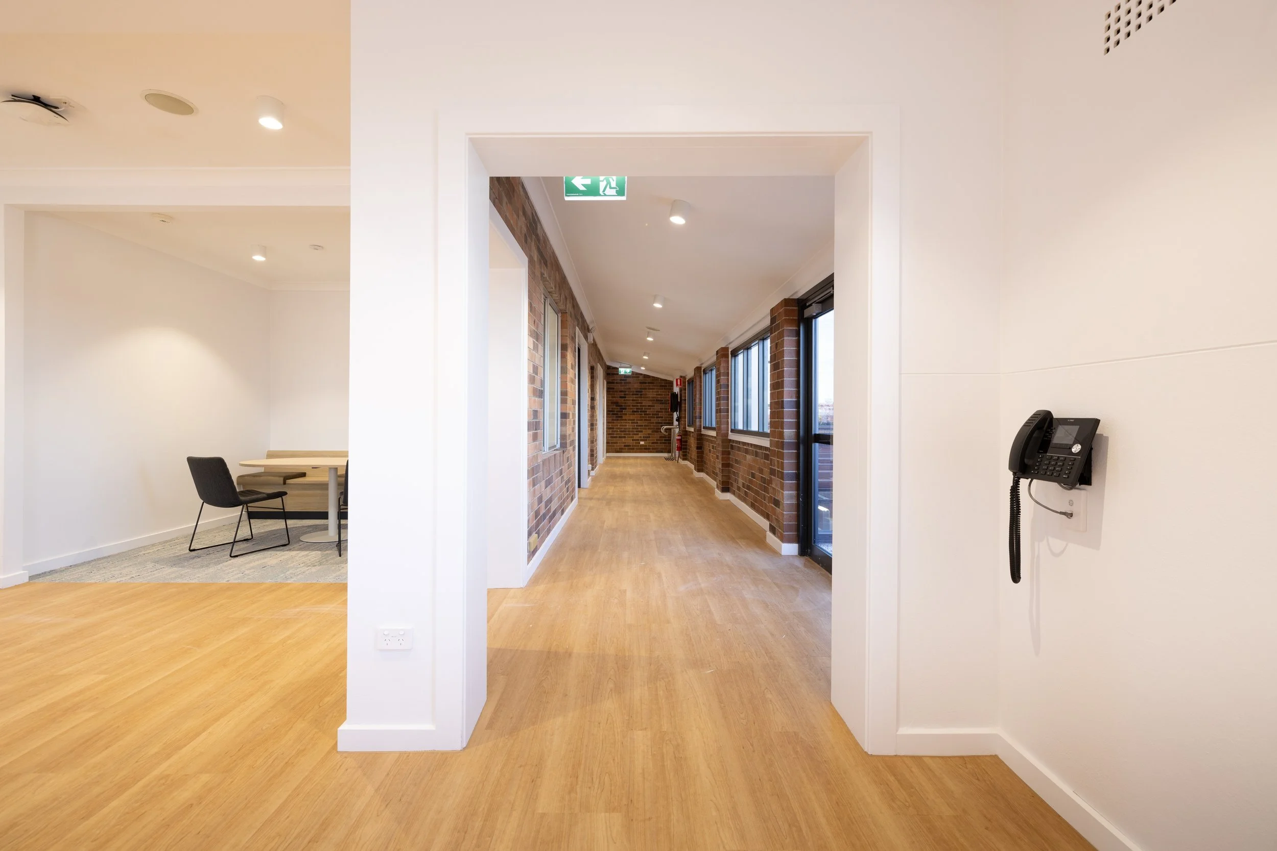 Empty hallway with brick walls and large windows, wooden floor, white walls, and a wall-mounted telephone.