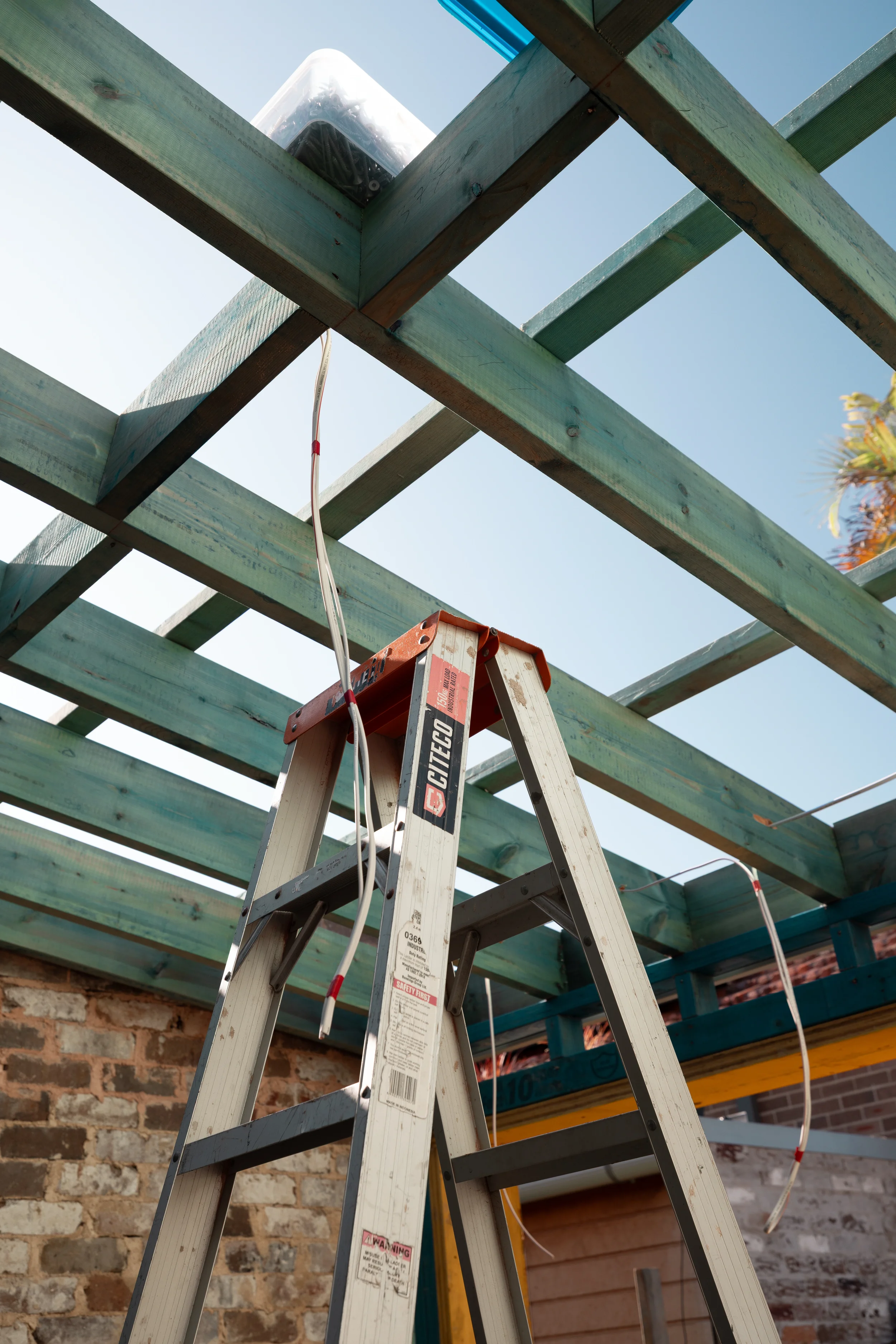 A metal stepladder positioned under a wooden porch structure that is under construction. The porch has turquoise wooden beams, and a large clear plastic container is placed on top of the porch framework. The background includes a brick wall and a brick building.