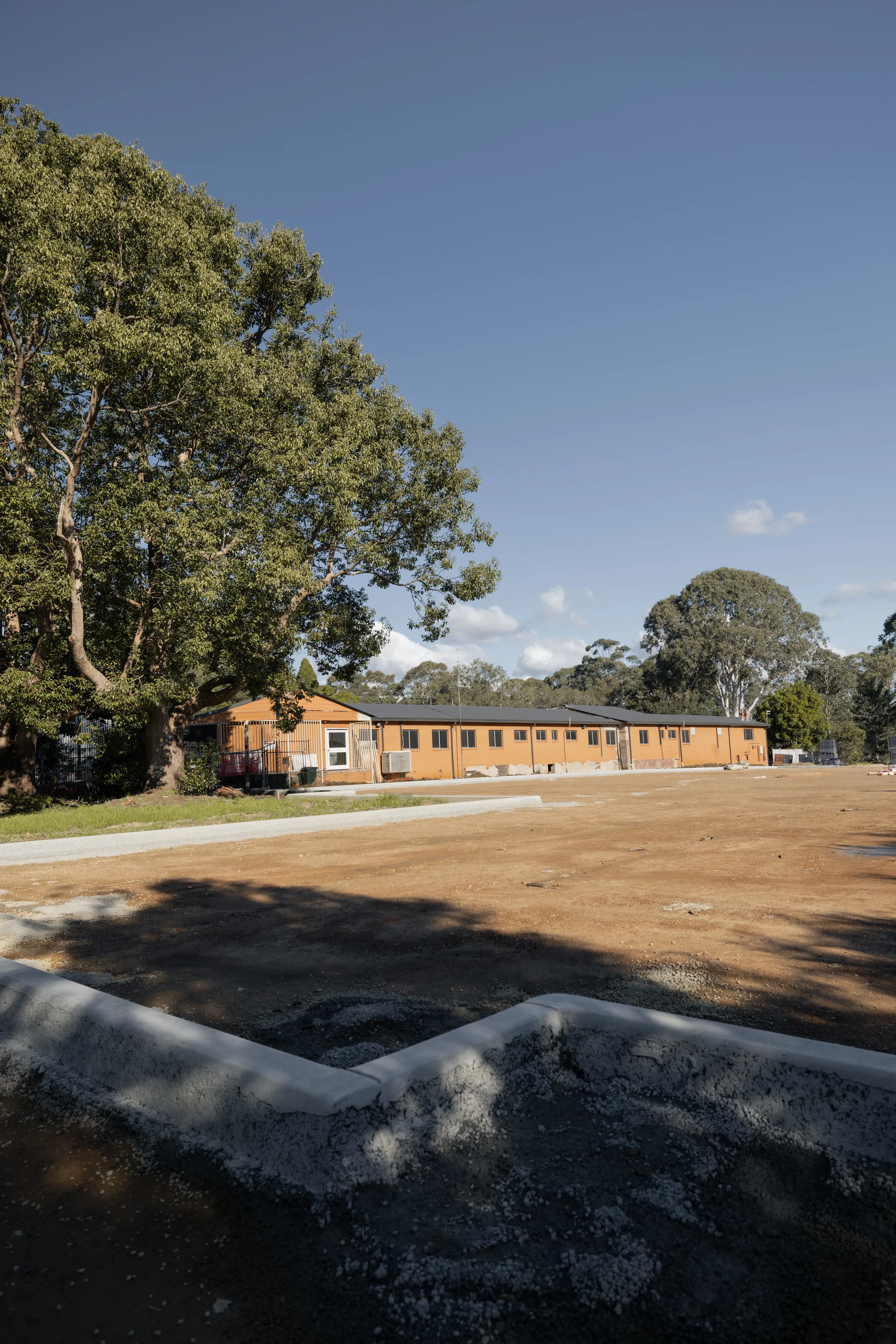 A newly constructed single-story brick building with large windows, surrounded by an open yard with some construction materials and areas of dirt and gravel. Several tall trees are in the background under a blue sky with a few clouds.