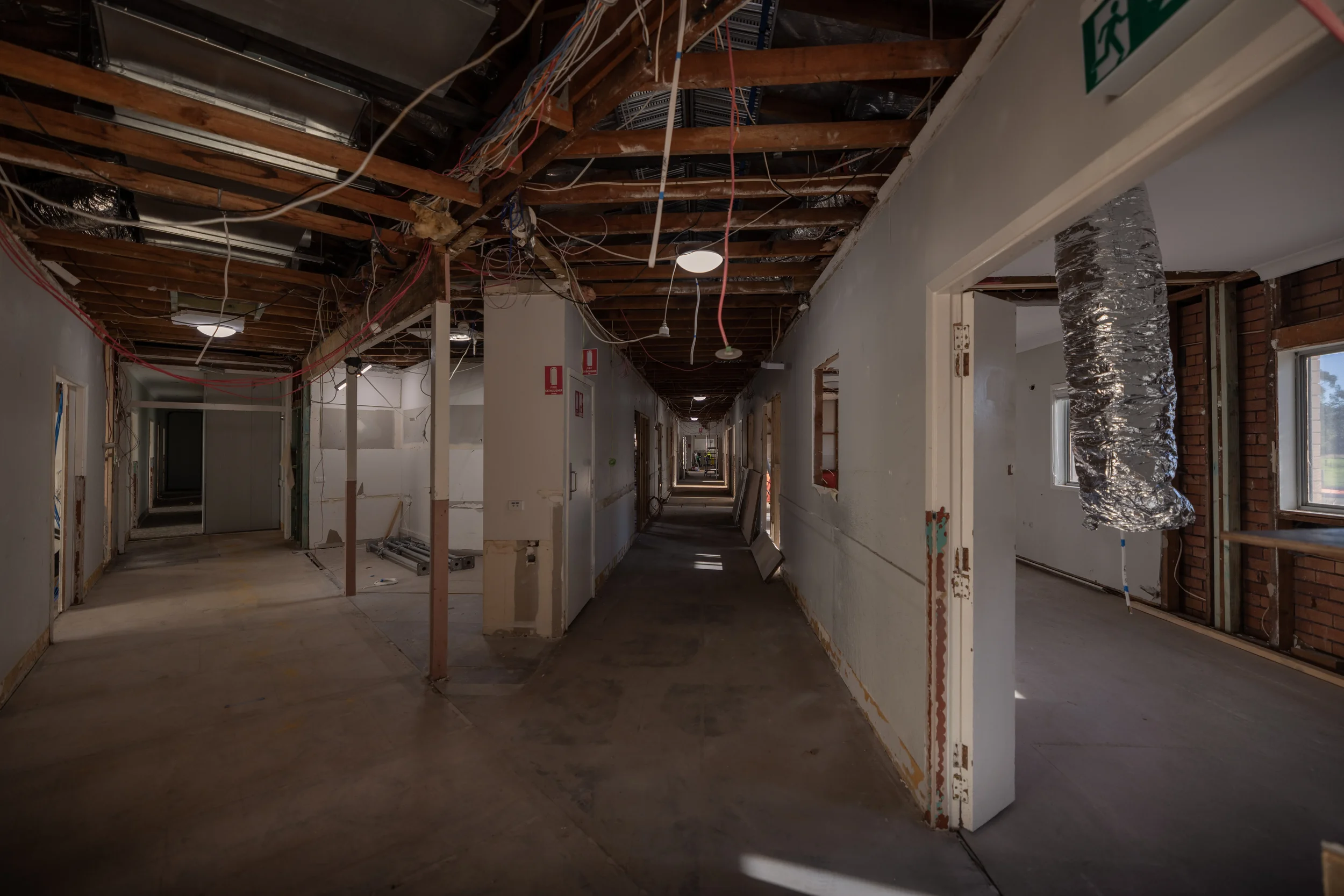 Construction site of an interior hallway with exposed wooden beams, wiring, and ventilation ductwork, with open doorways on either side.