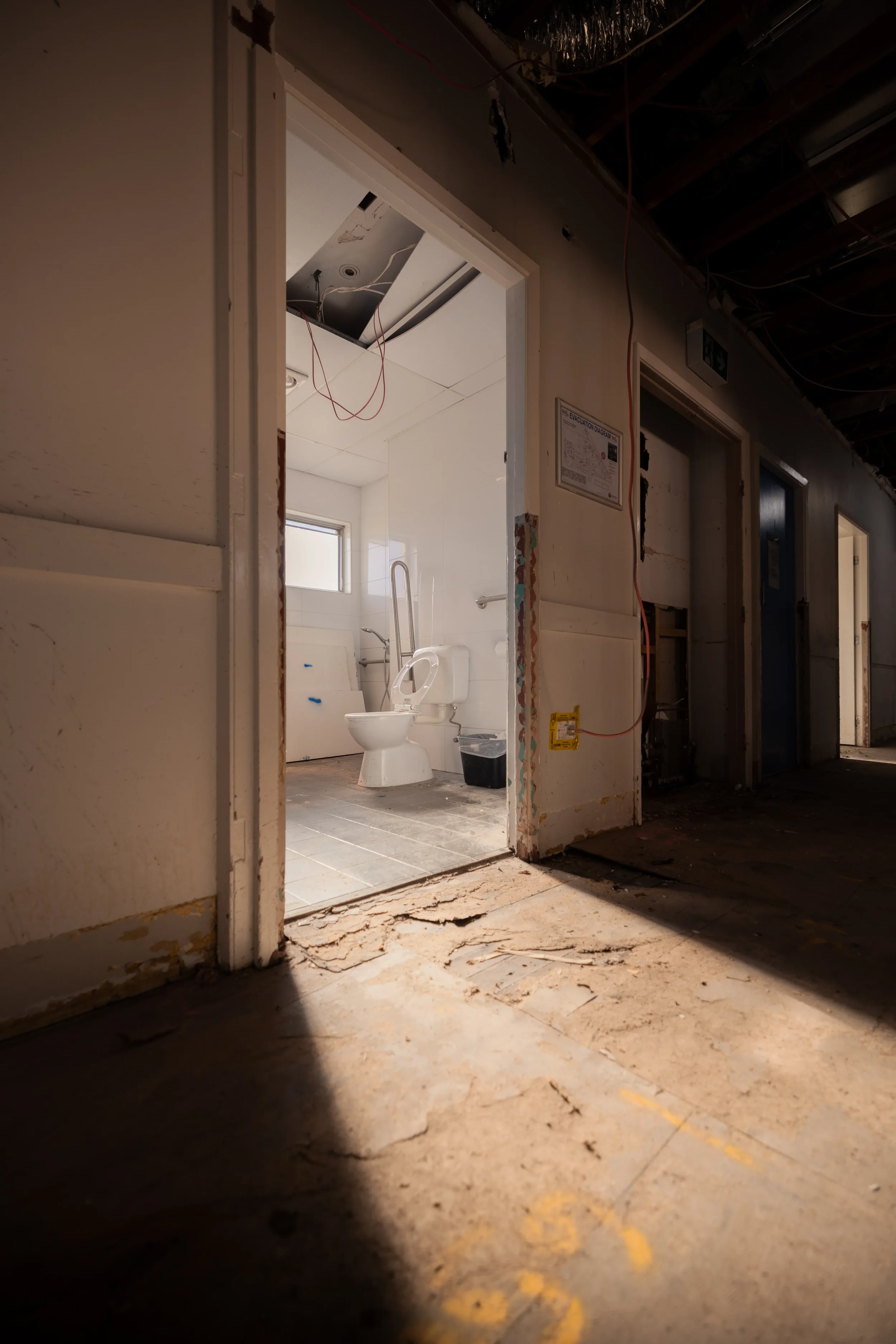 Interior of a building under renovation, showing a partially demolished bathroom with a toilet, tiled walls, and exposed ceiling with wires, viewed through a doorway with damaged floor tiles.