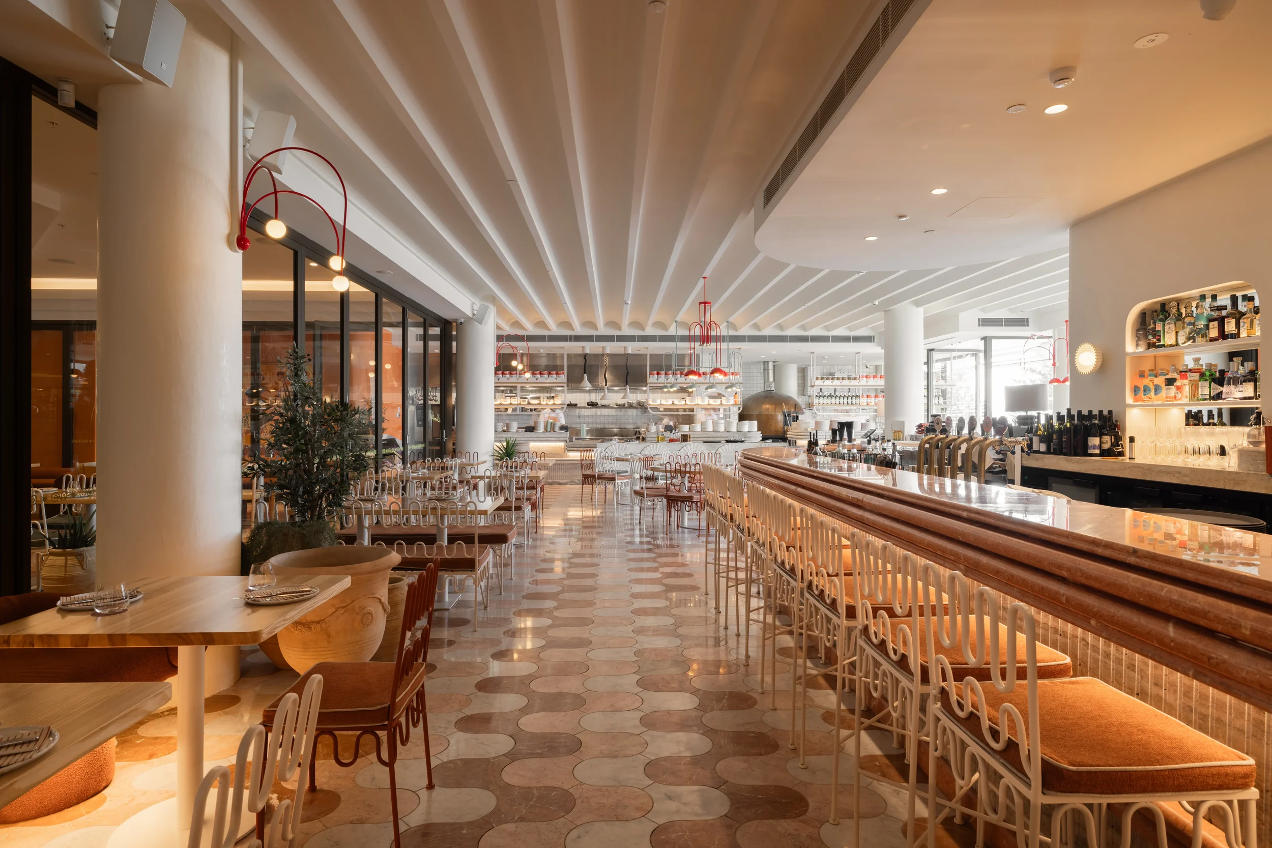 Interior view of a modern restaurant with white and orange seating, a marble bar counter, hanging red light fixtures, and a kitchen area in the background.