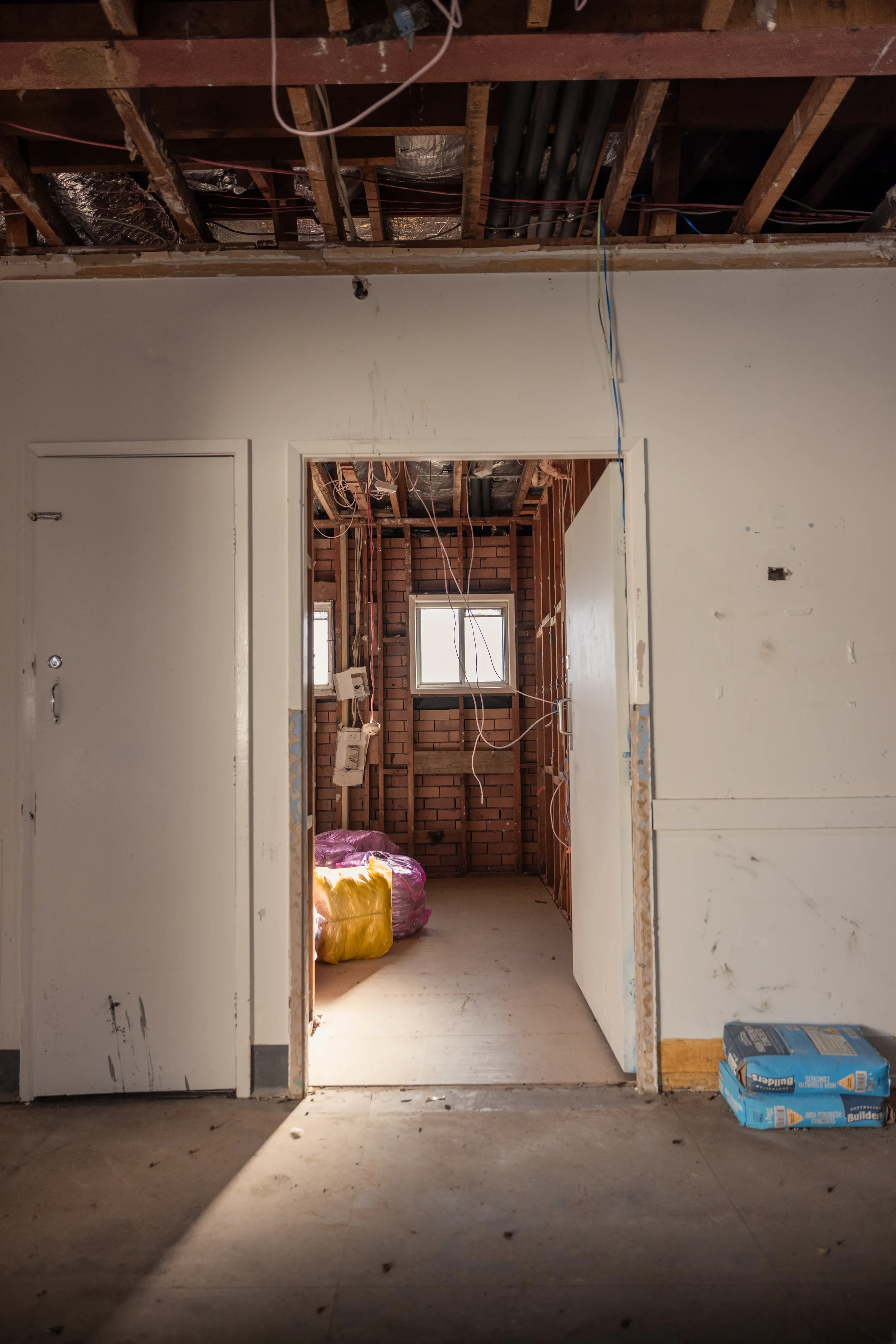 Interior view of a room under renovation with exposed ceiling showing wooden beams, pipes, and wires; doorway leading to another room with unfinished walls, windows, and construction supplies including insulation and a box of drywall screws.