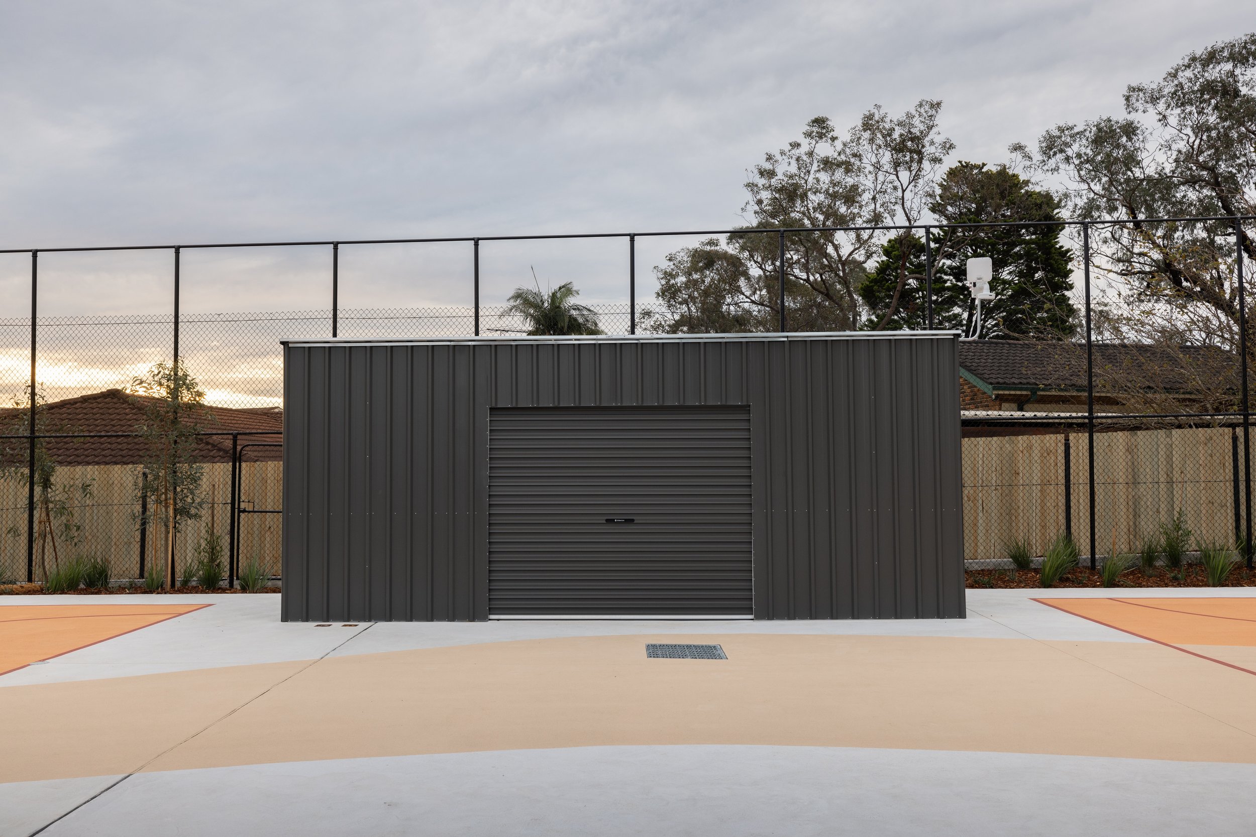 A small black metal shed with a roll-up door, situated on a concrete surface with a fenced backyard and trees in the background.