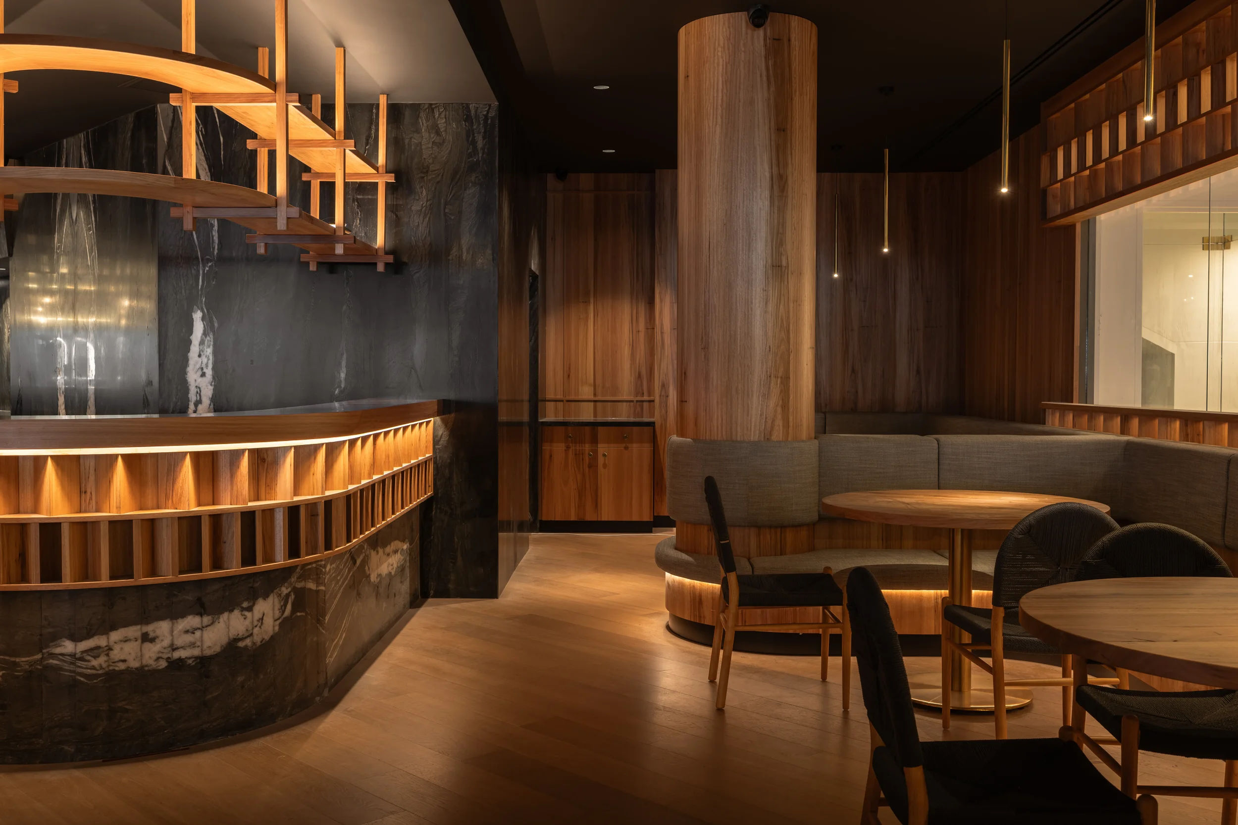 Interior of a modern restaurant with a curved wooden bar, black marble walls, wooden columns, and round tables with chairs, illuminated by pendant lights.