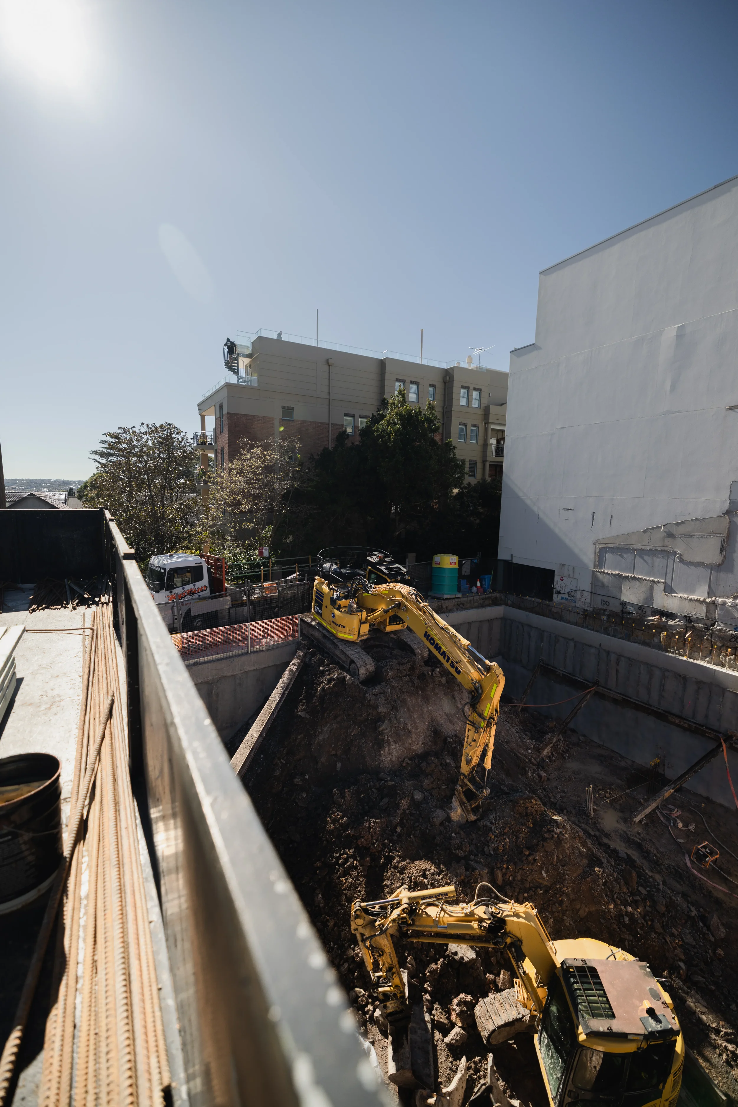 Construction site with excavator digging a deep trench, apartment buildings in the background, and a clear blue sky.