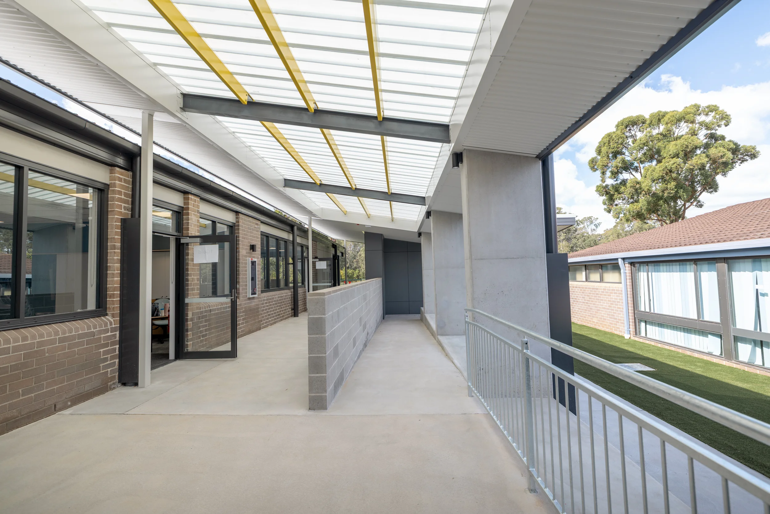 Exterior view of a modern building with brick and concrete walls, large windows, a glass door, and a covered walkway with a metal railing. The roof has transparent panels allowing natural light, and there are trees and a house with a tiled roof in th
