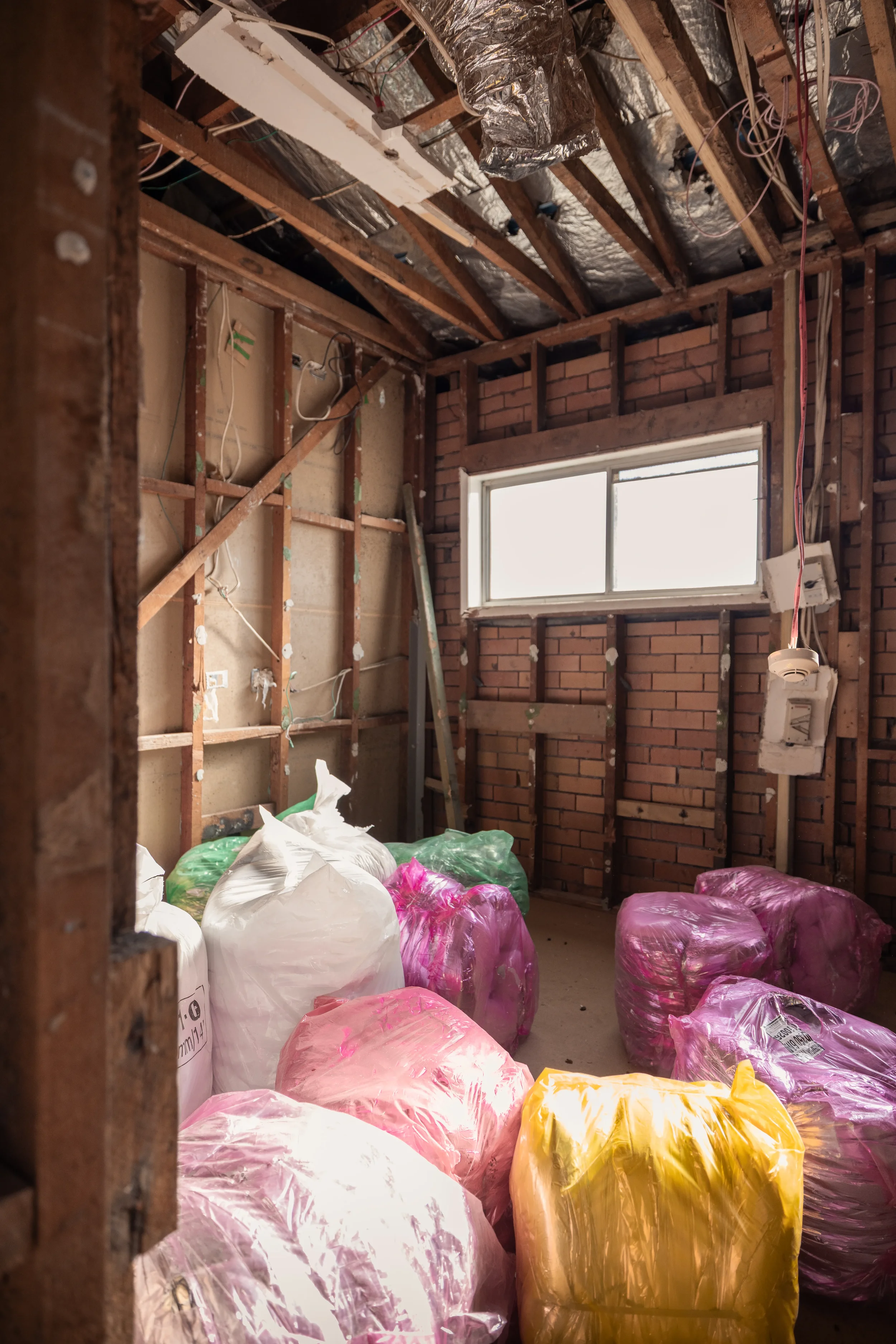 Room under renovation with exposed wooden beams, electrical wiring, and insulation material. Several large plastic bags filled with material are on the floor, with a window on the brick wall.
