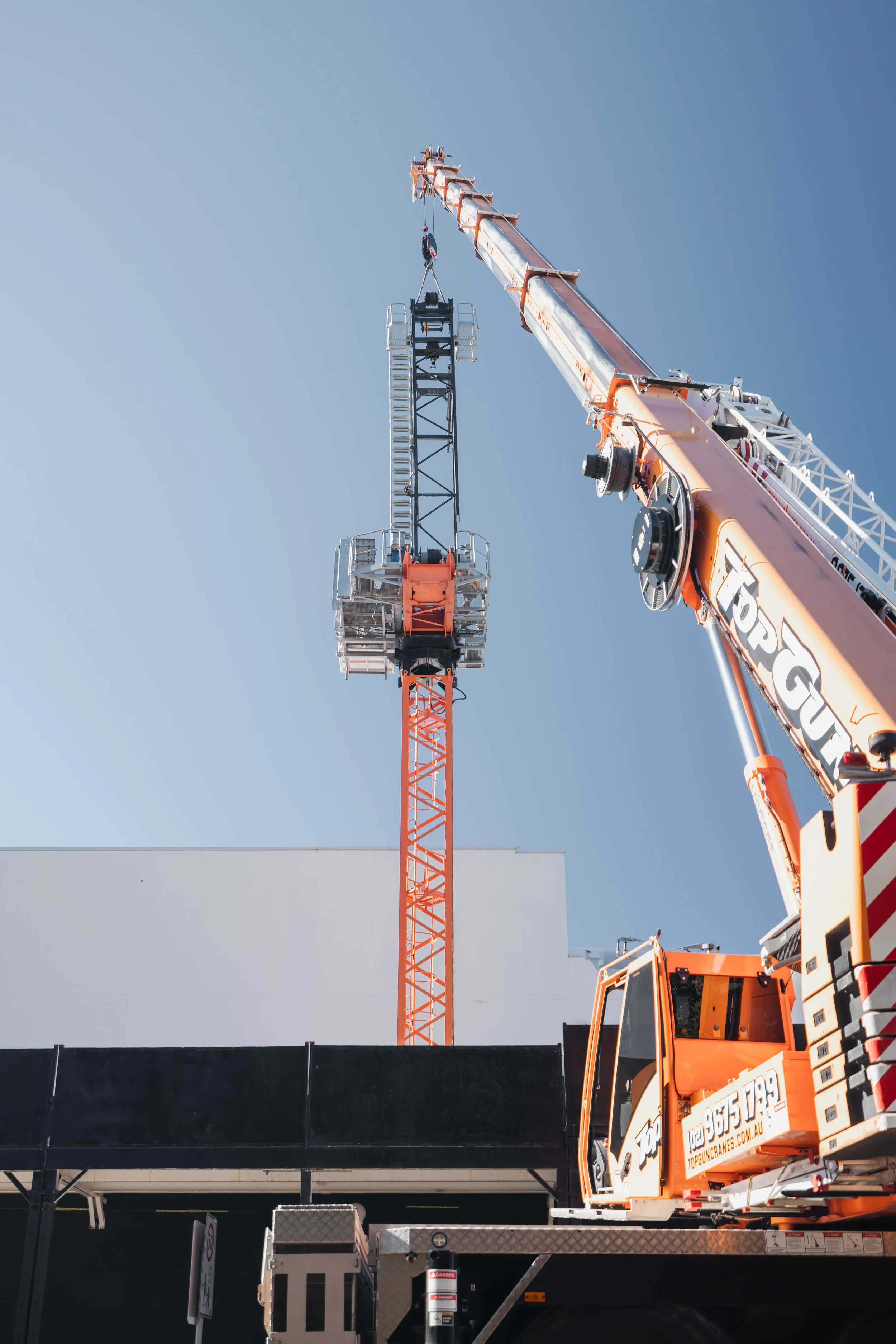 A large orange and white mobile crane lifting a black structure with a metal ladder against a clear blue sky.