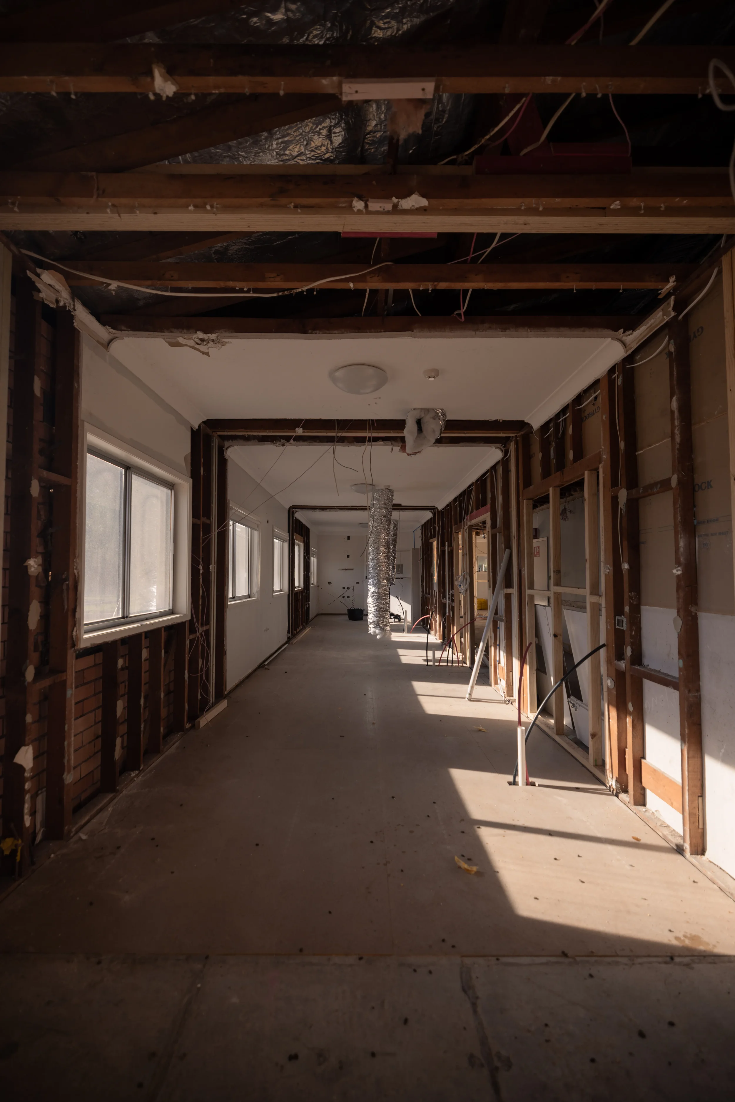 An interior space under renovation with exposed wooden framing, ceiling insulation, and unfinished walls, showing multiple windows along the side and construction debris on the floor.