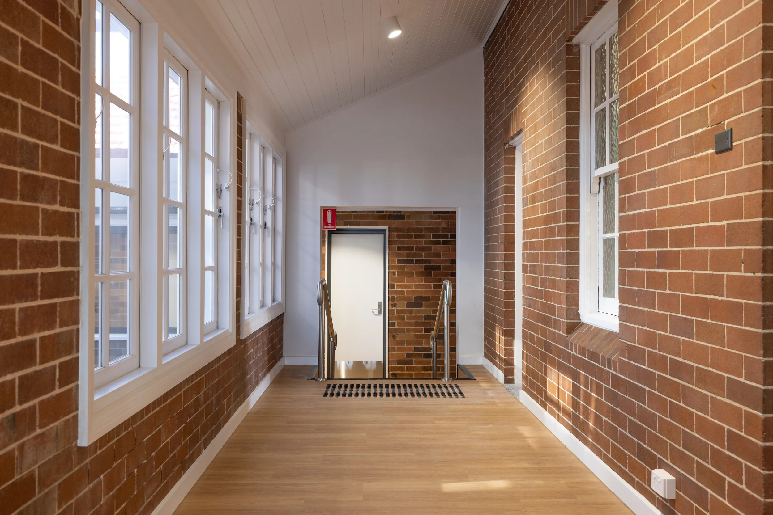 Interior hallway with brick walls on both sides, large windows on the left, a staircase at the end, a fire extinguisher on the wall, and ceiling lights.