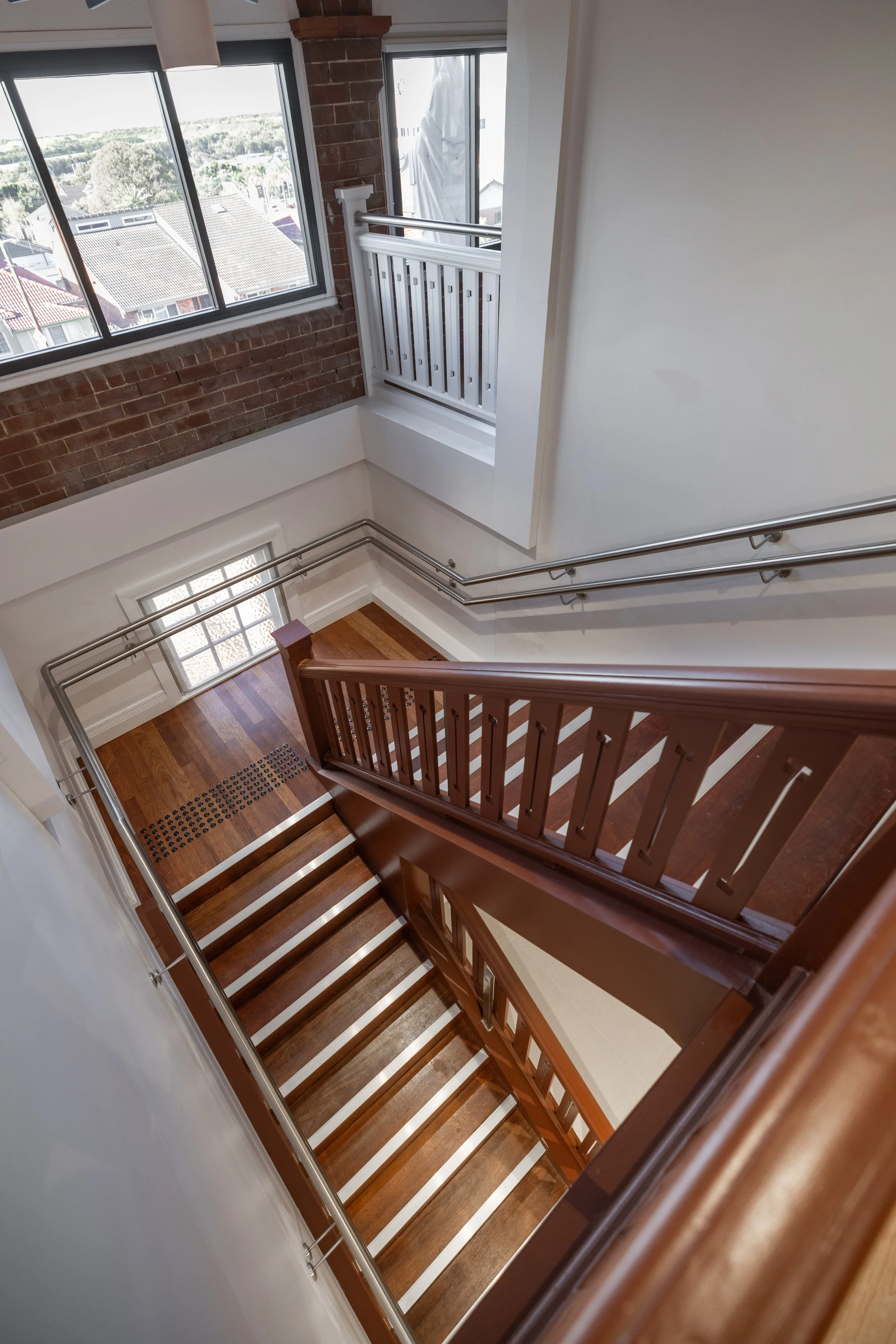 View looking down at a staircase with wooden steps and brown handrails in a well-lit house, with large windows showing neighboring houses and trees outside.