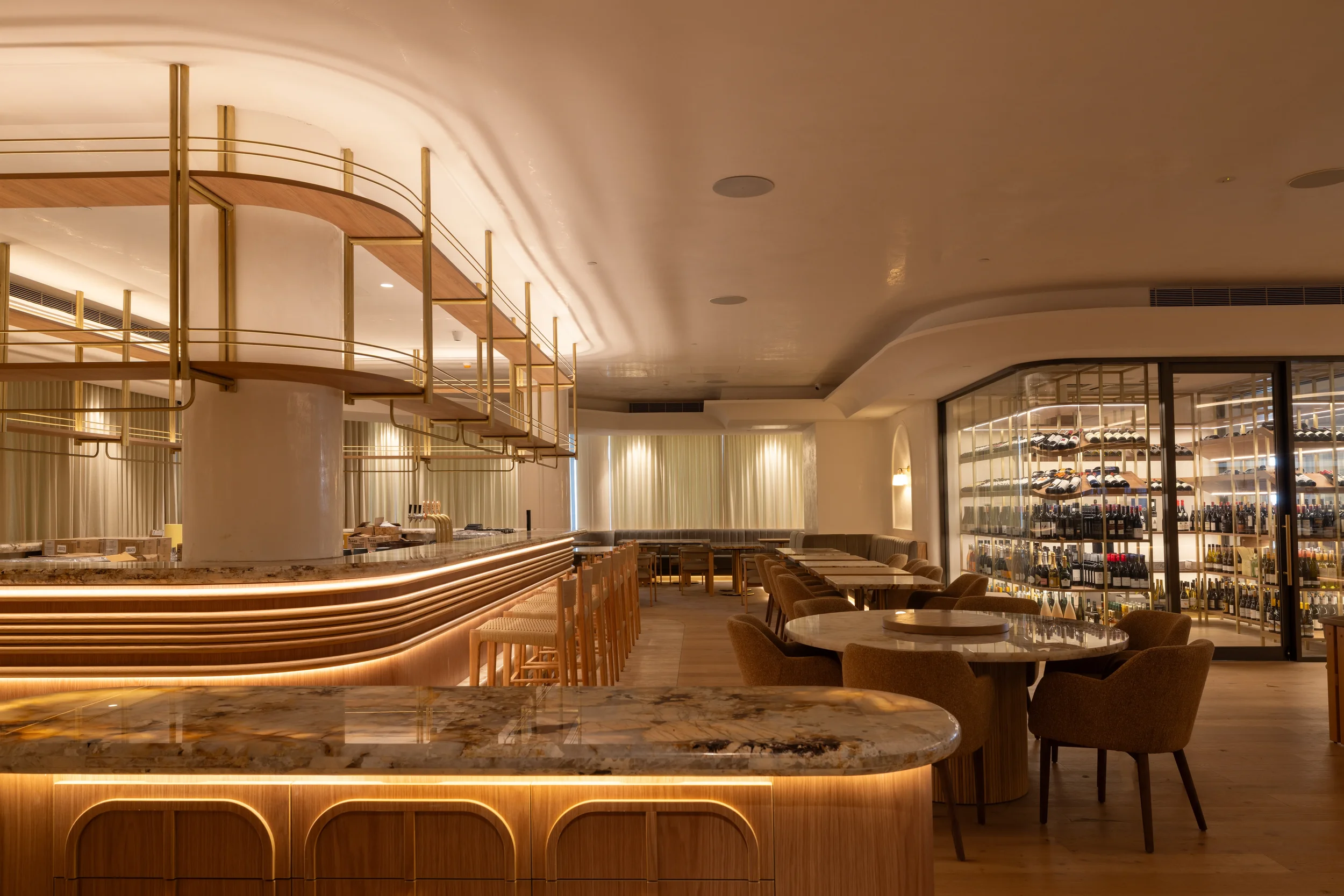 Interior of a modern upscale restaurant or bar with a curved marble counter, bar stools, and wooden shelving with bottle displays, warm lighting, and upholstered chairs around tables.