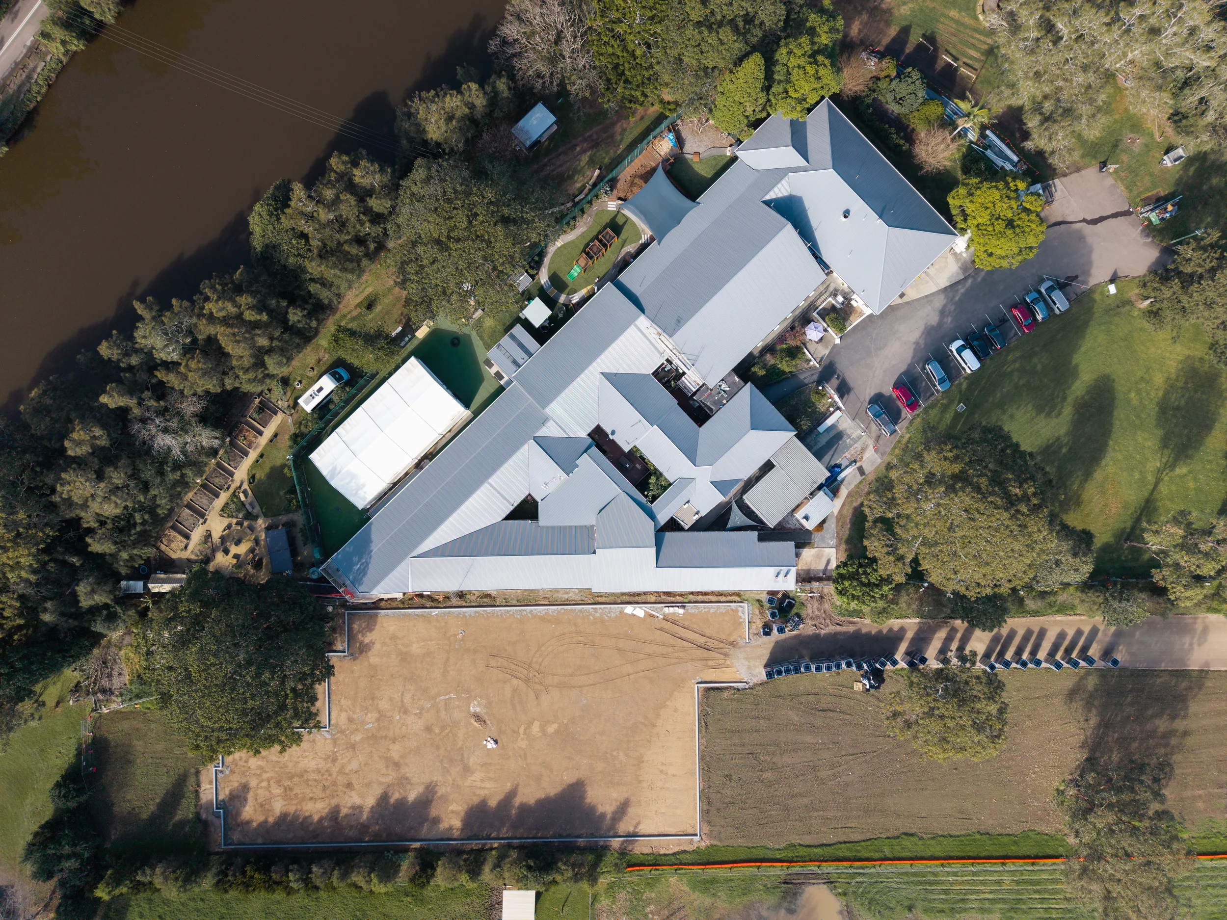 Aerial view of a large building complex with a gray roof, surrounded by green trees, some playground areas, parking lots with several cars, and a water body on the left side of the image.