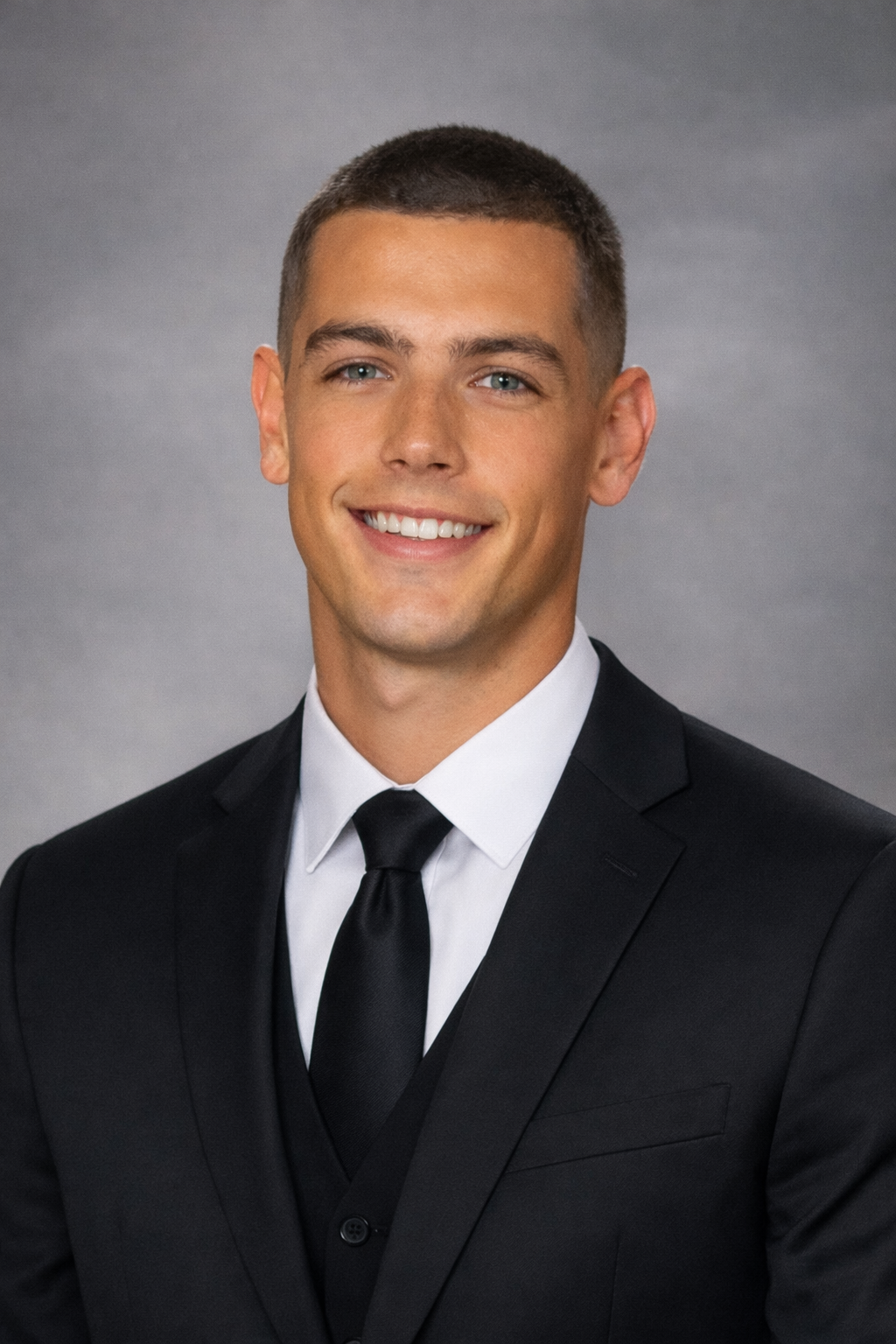 A man with dark, curly hair, glasses, and a beard looking off to the side, wearing a dark green sweater, sitting indoors with a plant in the background.