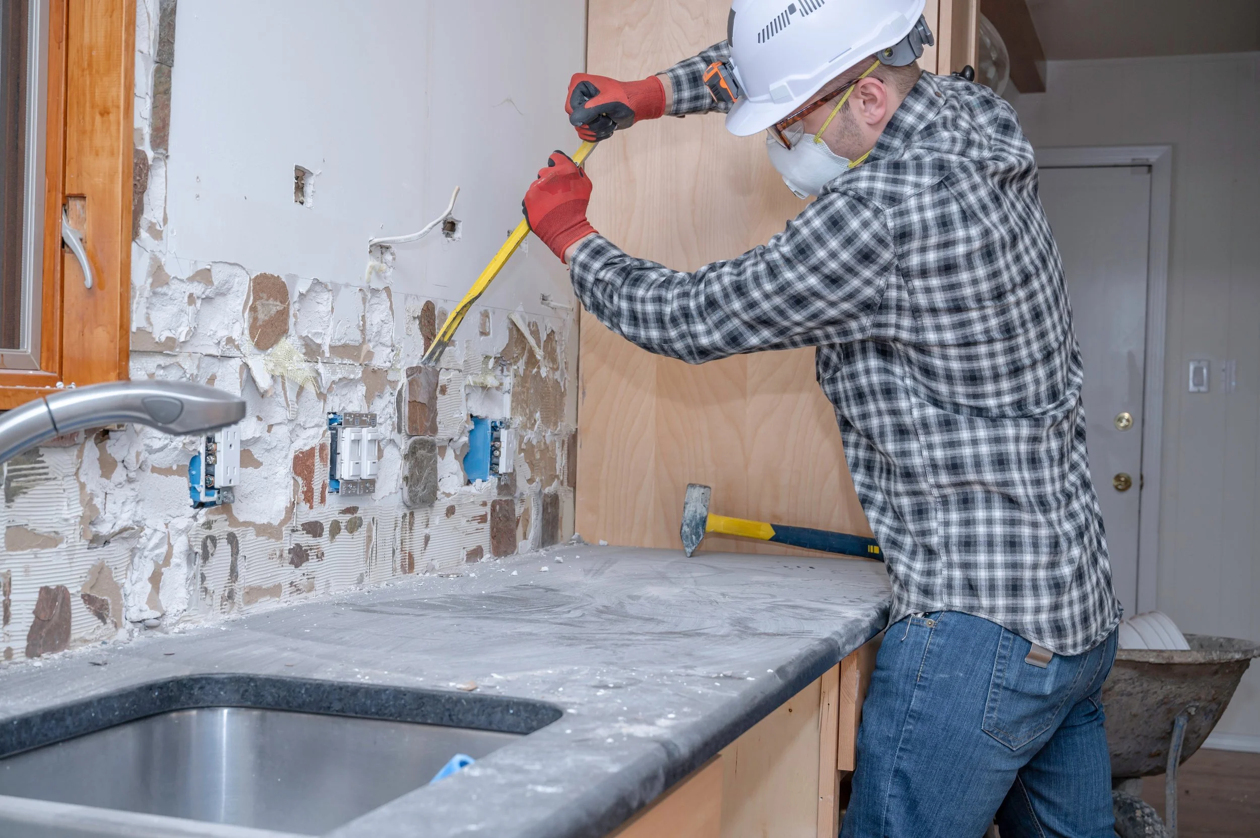 A worker wearing a hard hat, face mask, glasses, plaid shirt, and gloves uses a crowbar to remove drywall from a wall in a kitchen renovation.