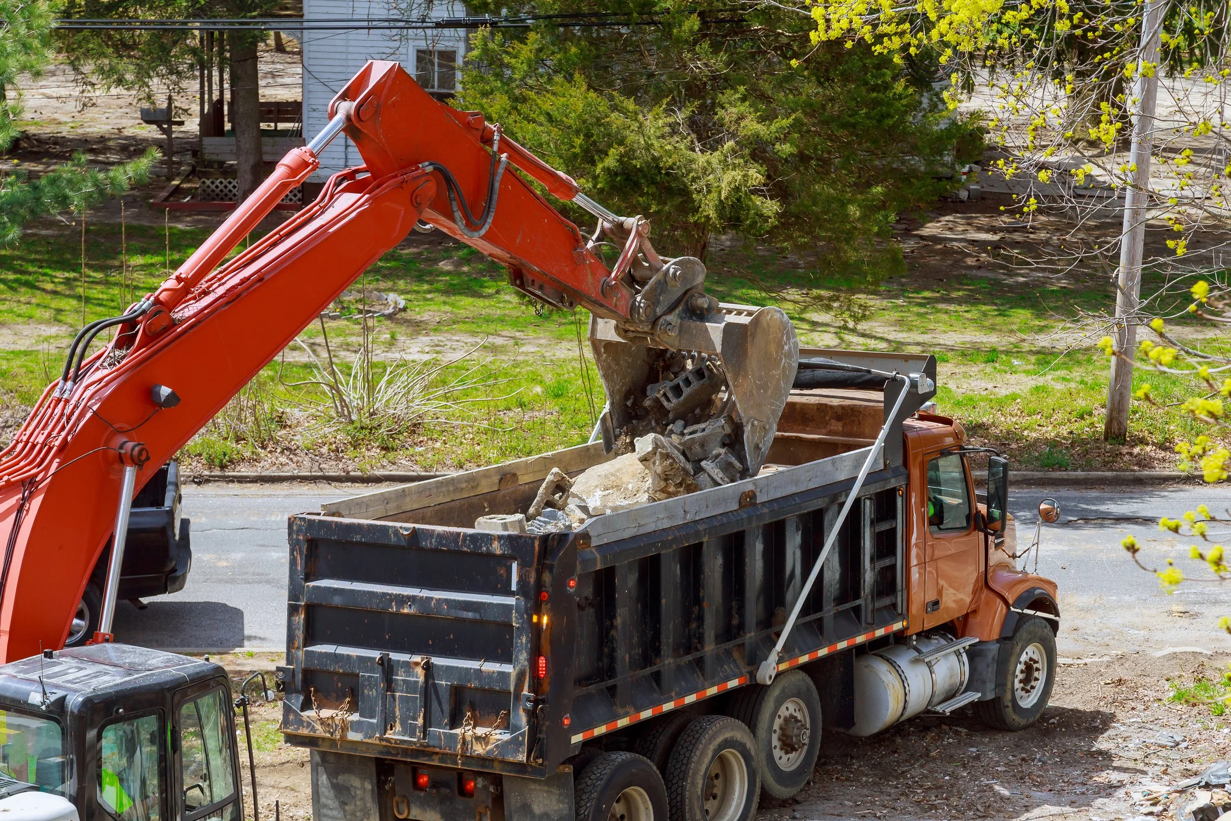 A large orange excavator loading rocks into a black dump truck on a residential street with trees and grass.