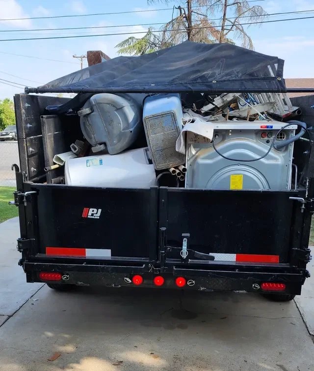 A trailer filled with discarded household appliances and trash, including a washing machine, dryer, large metal container, and other debris, parked on a concrete driveway.