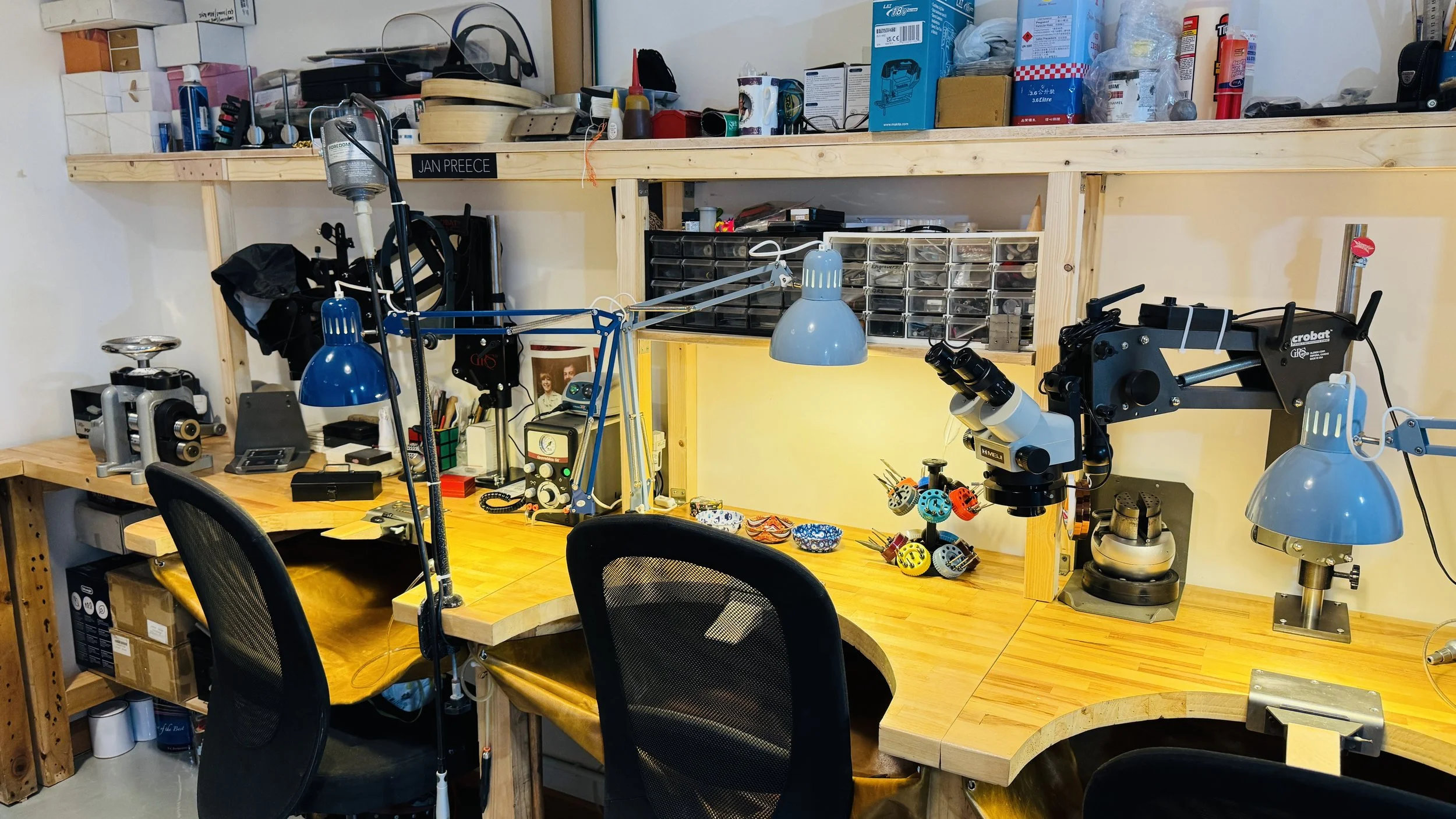 A woodworking or electronics workbench with various tools, microscopes, and equipment, against a wall with shelves holding boxes and supplies, and illuminated by multiple desk lamps.