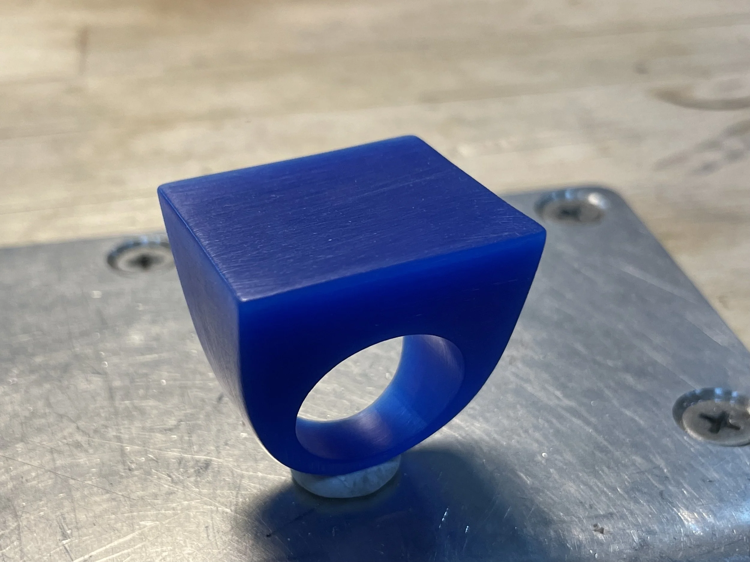 Close-up of a blue, rectangular object with a textured surface, attached to a metal or plastic base with screws. The background features a wooden surface.