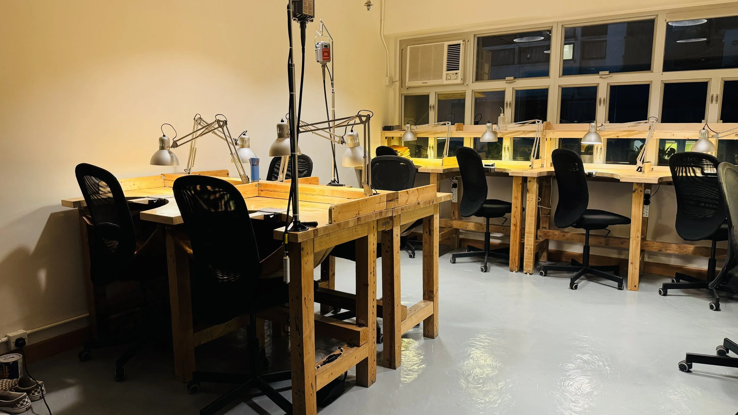 An empty coworking space or office with wooden desks, black mesh chairs, and adjustable desk lamps. The room has large windows with multiple panes and a window air conditioning unit. The lighting is warm, and personal items like notebooks and folders are on the desks.