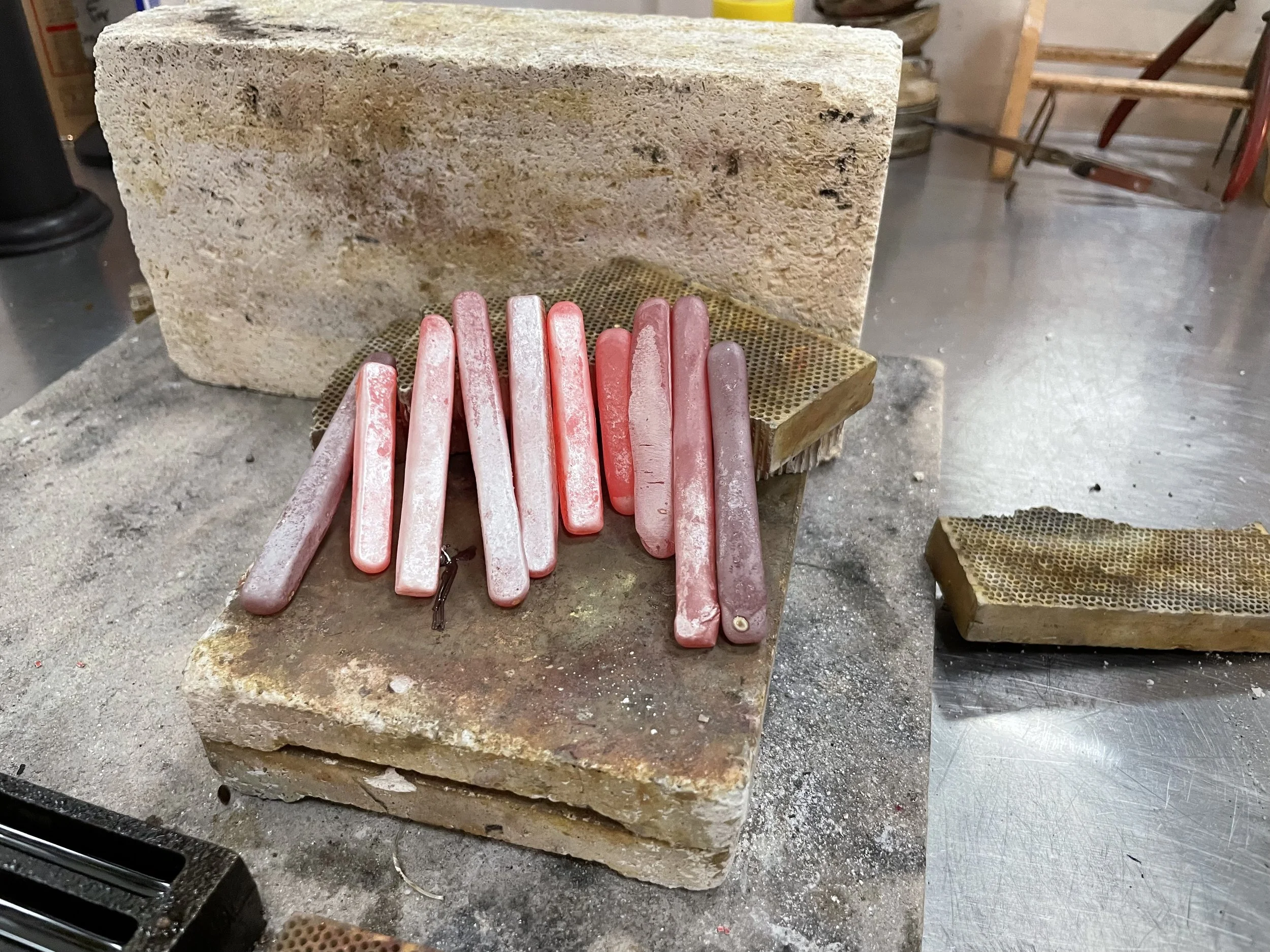 A large rectangular stone block with a rough surface, placed on a workbench. In front of the stone are several pink wax carving sticks, some with black stains. The workspace has a dusty surface with small debris, and tools and shelves can be seen in the background.