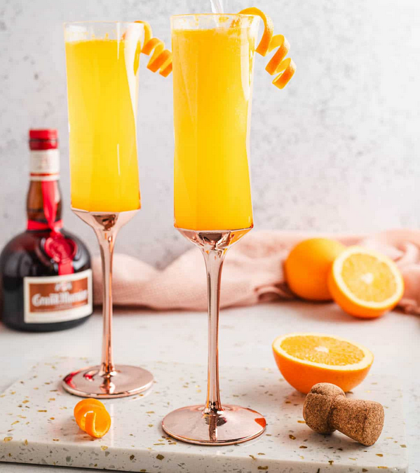 Two champagne glasses filled with orange yellow cocktails garnished with orange peel twists, alongside halved oranges, a bottle of Aperol, a pink cloth, and a cinnamon stick on a white surface.