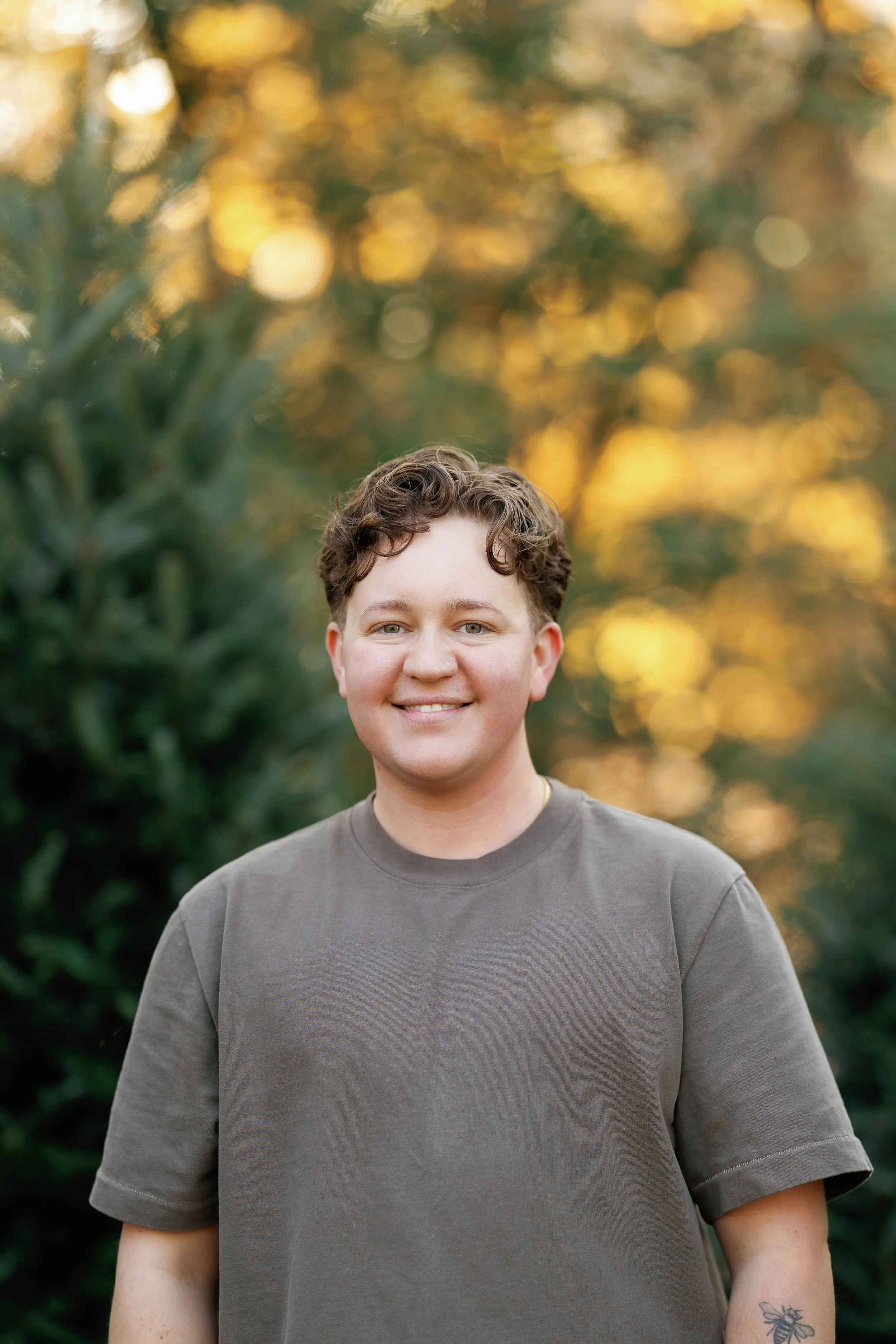 A young person with short, curly hair smiling outdoors during sunset, wearing a gray T-shirt, with blurred trees and warm golden light in the background.