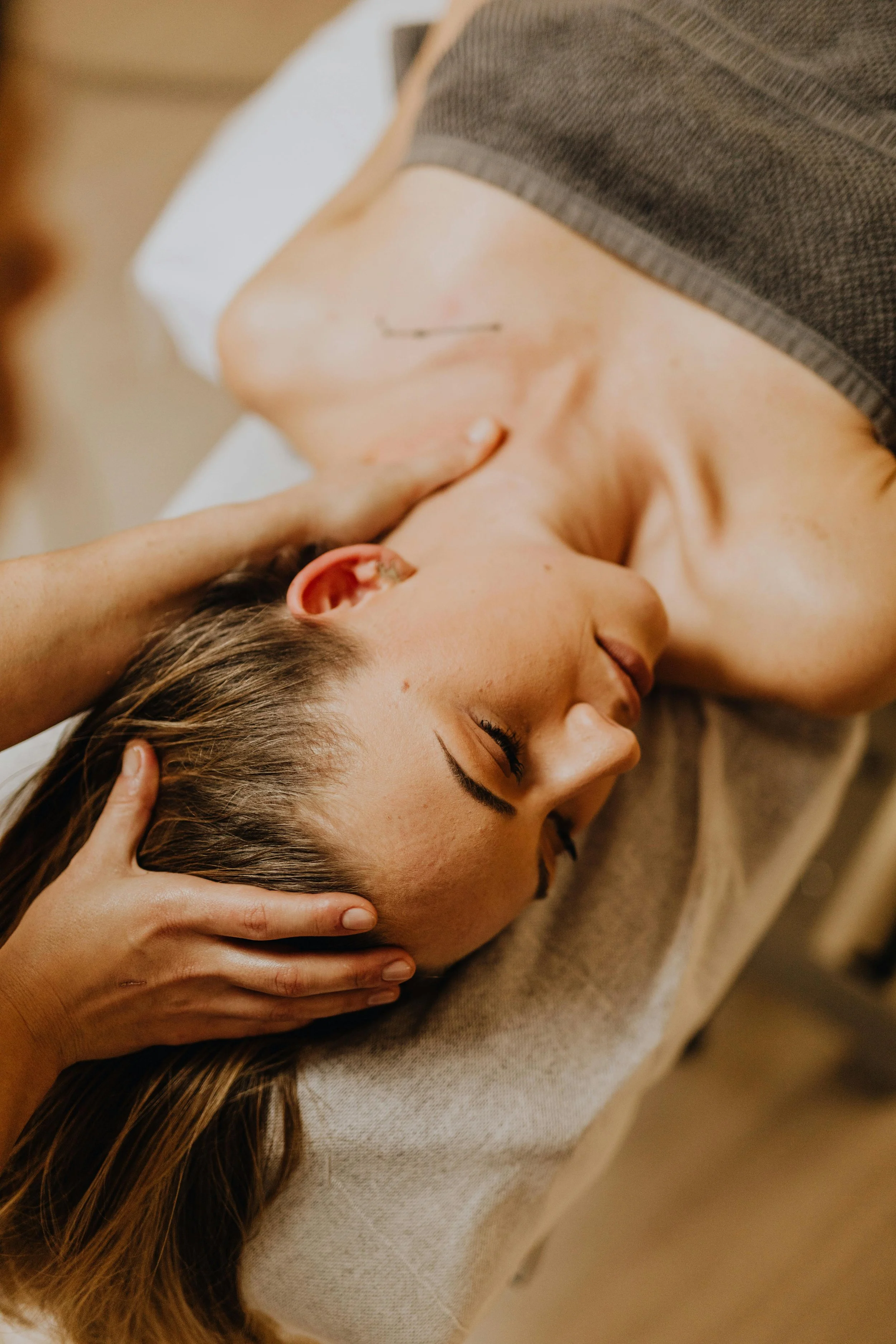 A young woman lying face-up on a massage table with her eyes closed, receiving a massage or therapy, with hands gently massaging her head.