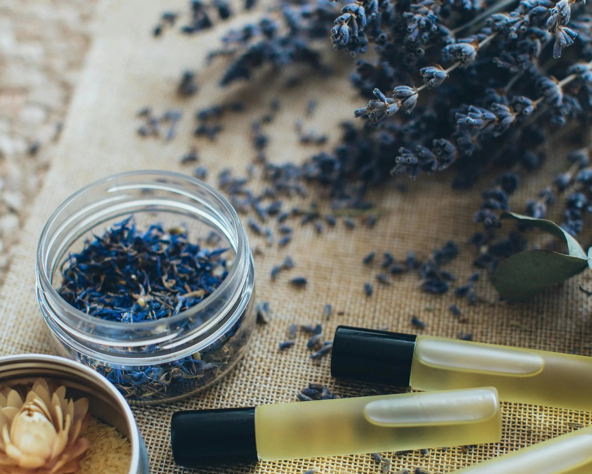 Dried lavender flowers in a glass jar, purple lavender bunches, two yellow roller bottles with black caps, a small bowl with a dried flower, and green leaves, all placed on a textured beige surface.