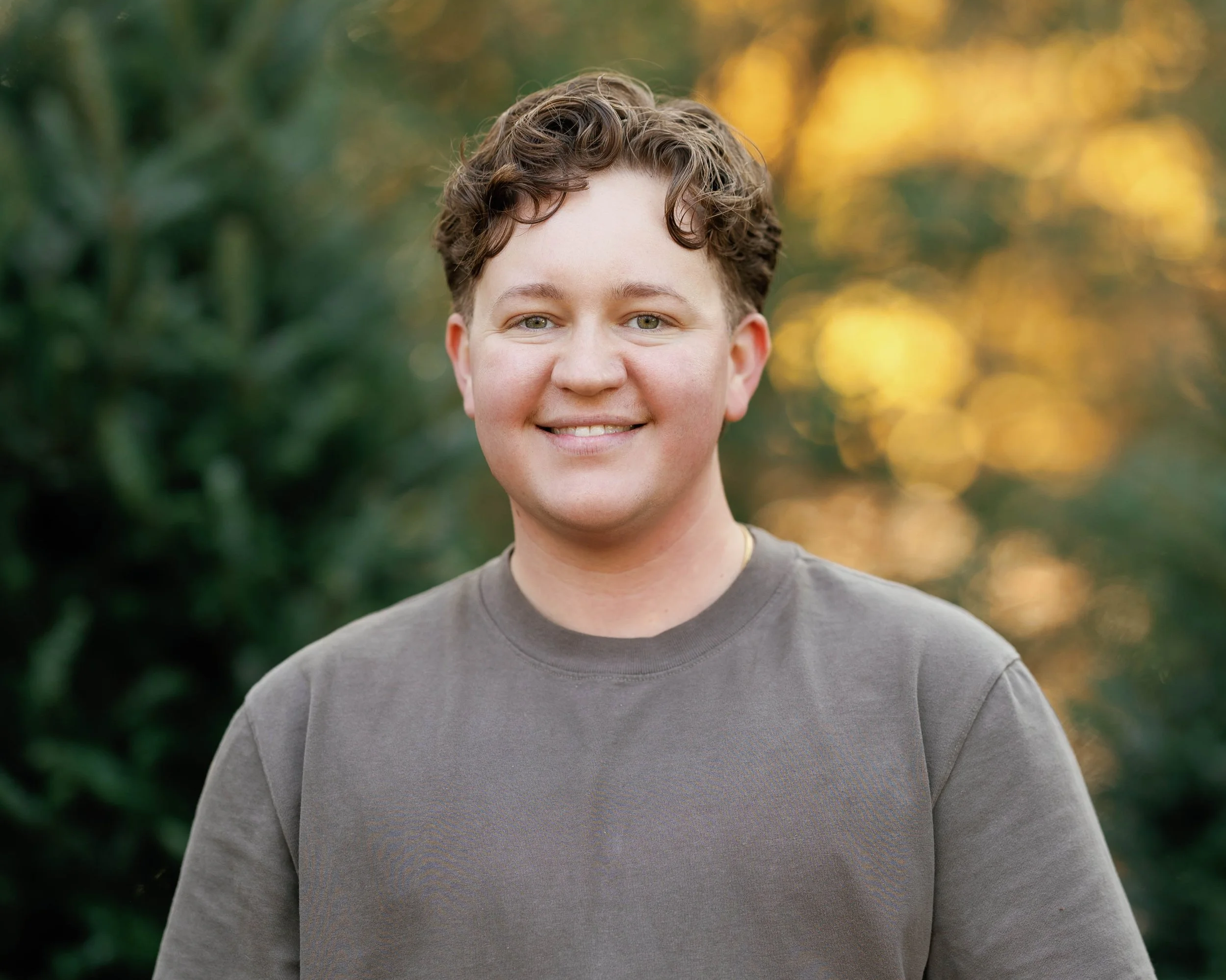 A young man with curly brown hair and a friendly smile standing outdoors with a blurred background of green trees and warm sunlight.
