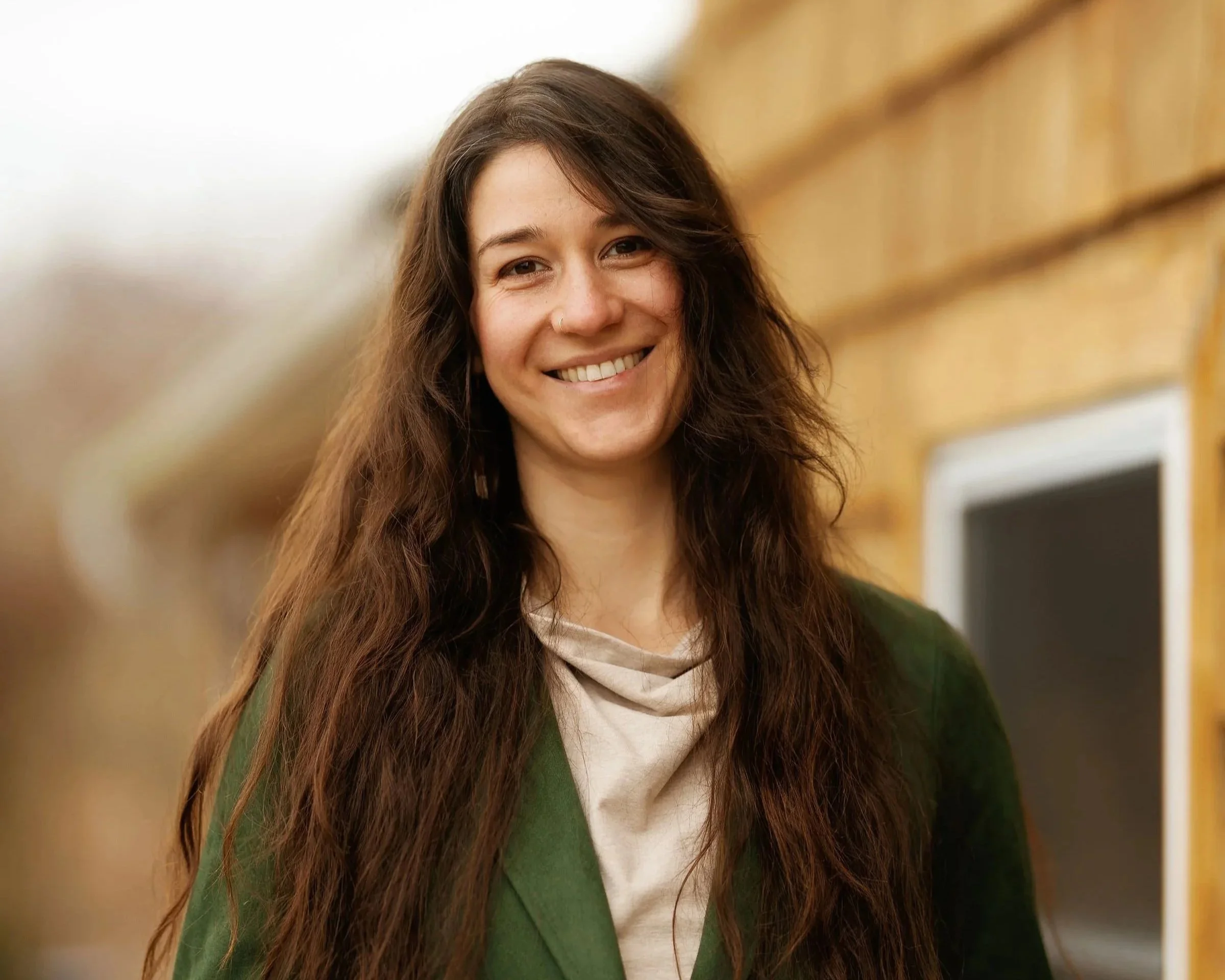A young woman with long wavy brown hair, wearing a beige sweater and colorful earrings, standing outside in front of a wooden house with a window and green plants.
