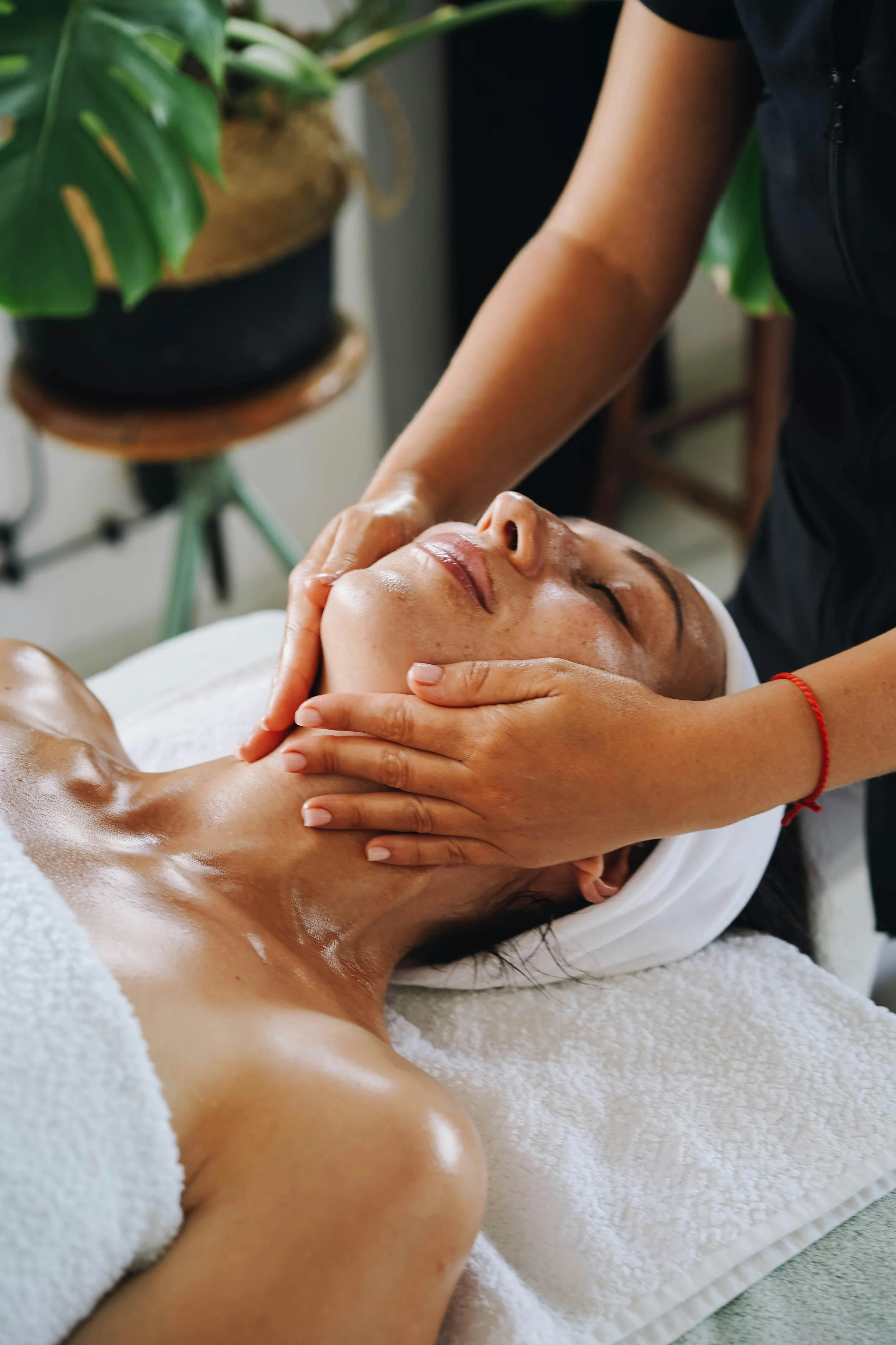 A woman receiving a facial massage at a spa, lying down with a towel wrapped around her body, while a therapist gently massages her face and neck.