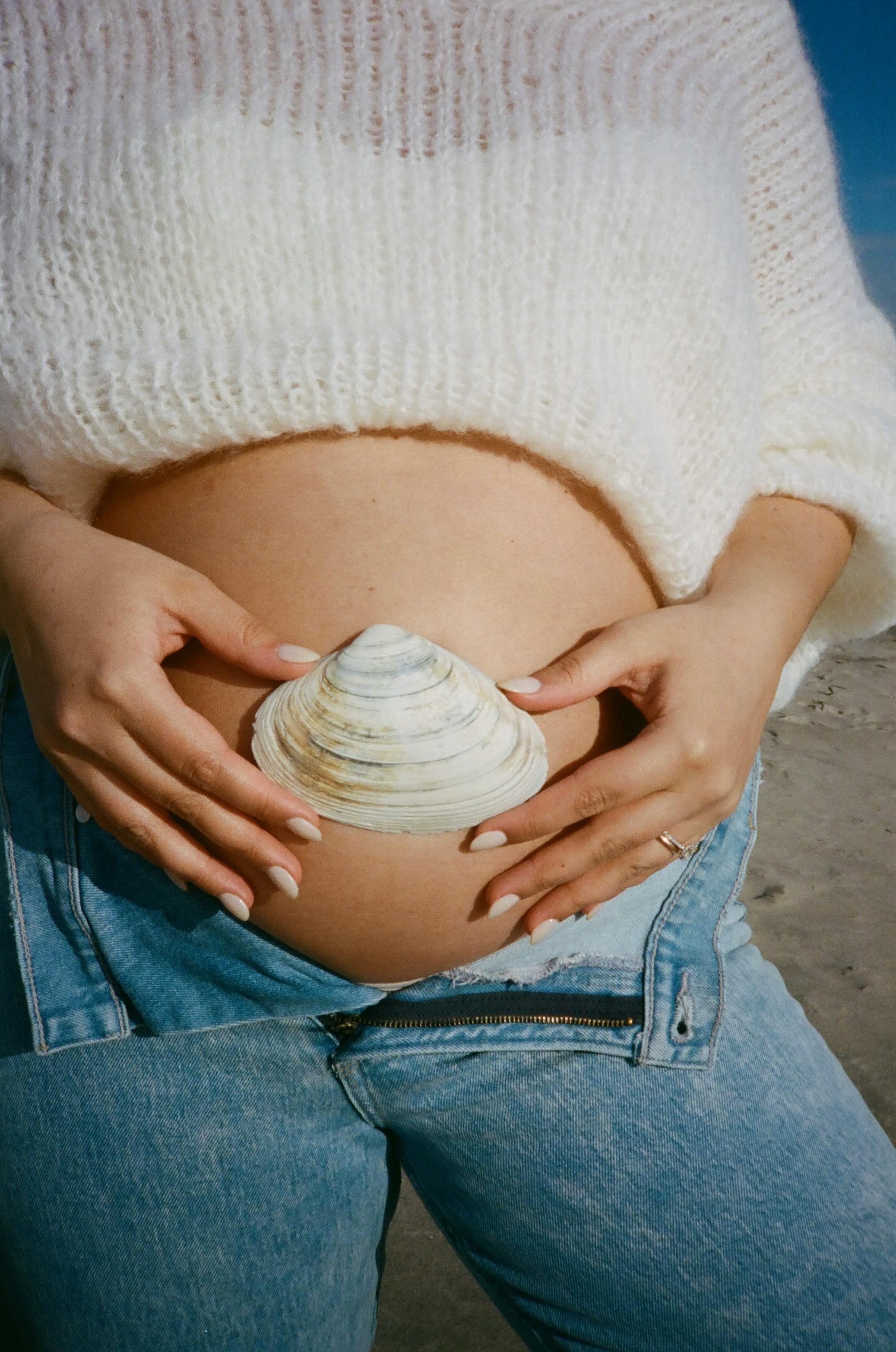 Close-up of a pregnant woman holding a seashell on her bare belly, wearing a white sweater and jeans, with beach sand visible in the background.