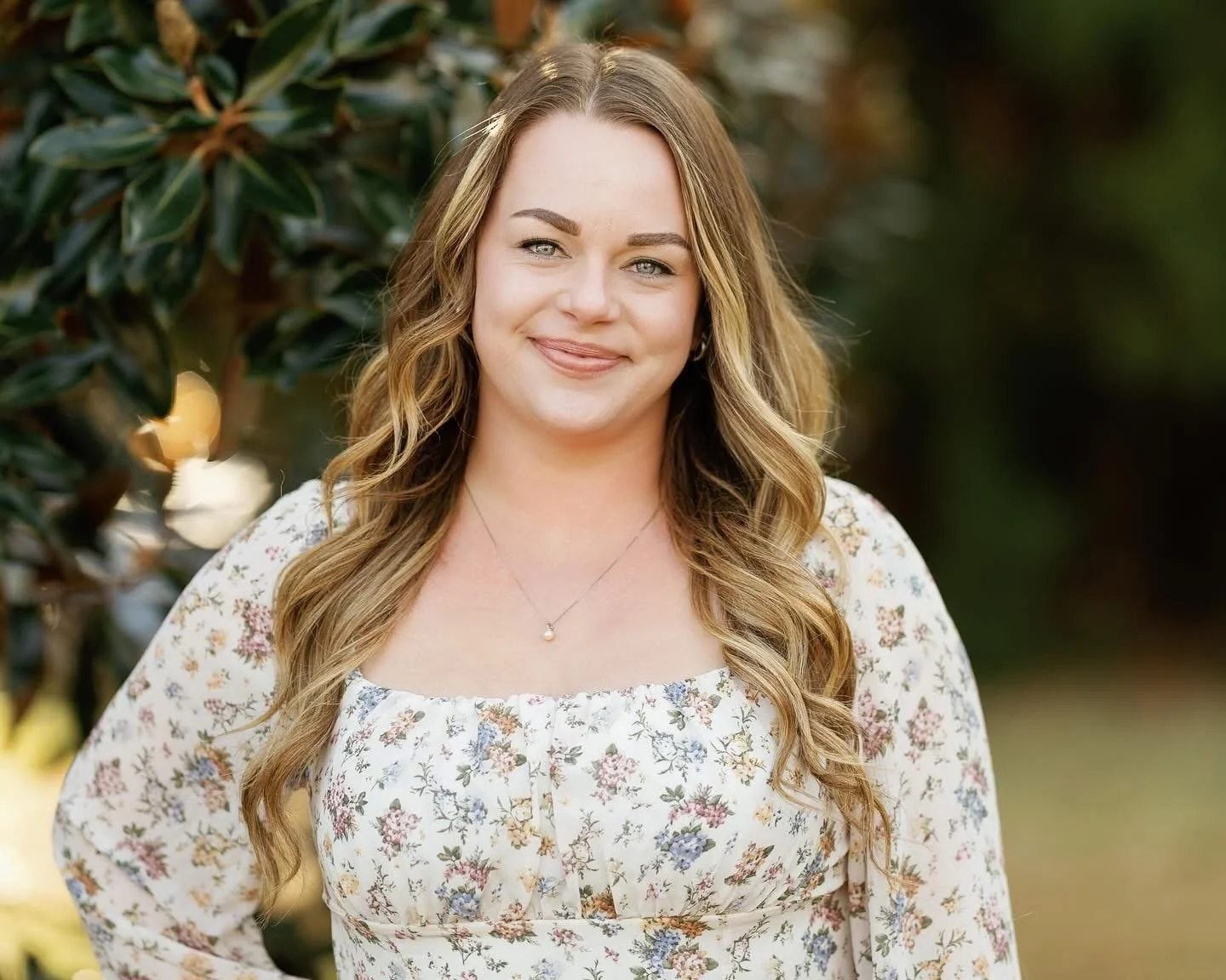 A young woman with long, wavy blonde hair and blue eyes, wearing a white floral blouse and a delicate pendant necklace, standing outdoors in front of green foliage.