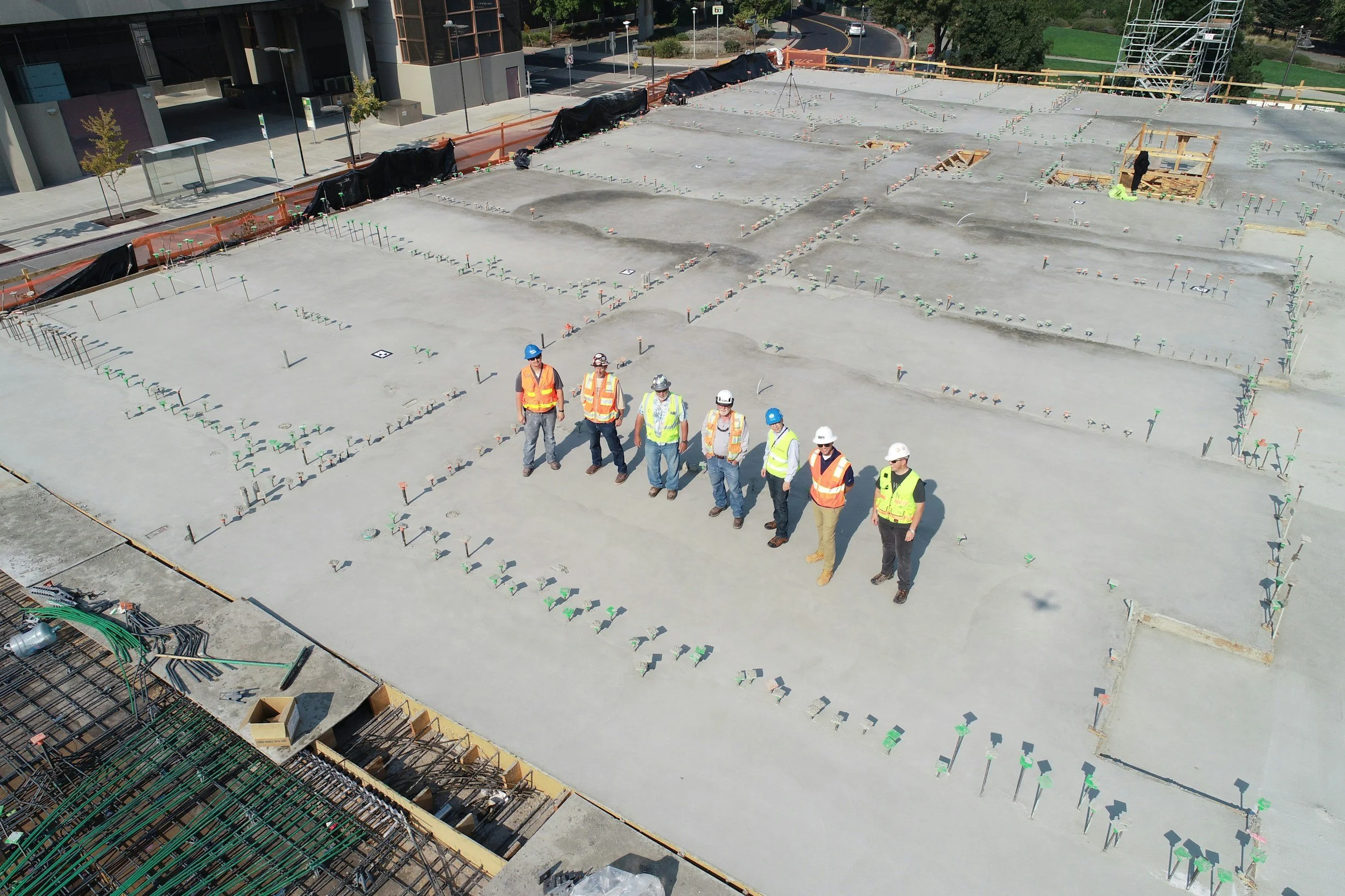 Construction workers in safety vests and helmets stand on freshly poured concrete at a building site. The site is organized with numerous small markers and rebar outlines for future construction.