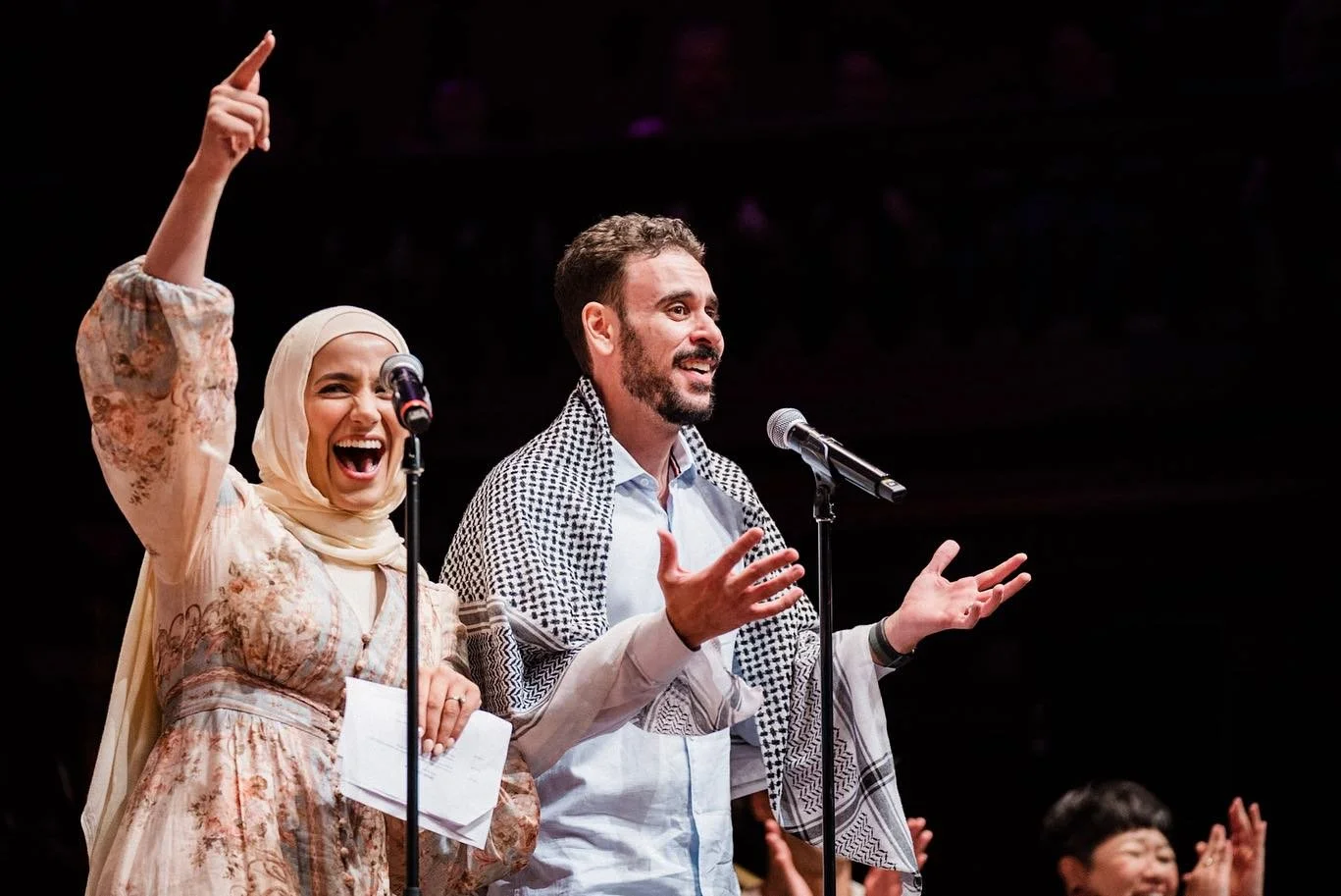 Two people speaking on stage; a woman wearing a hijab and a man with a beard, both smiling and gesturing.