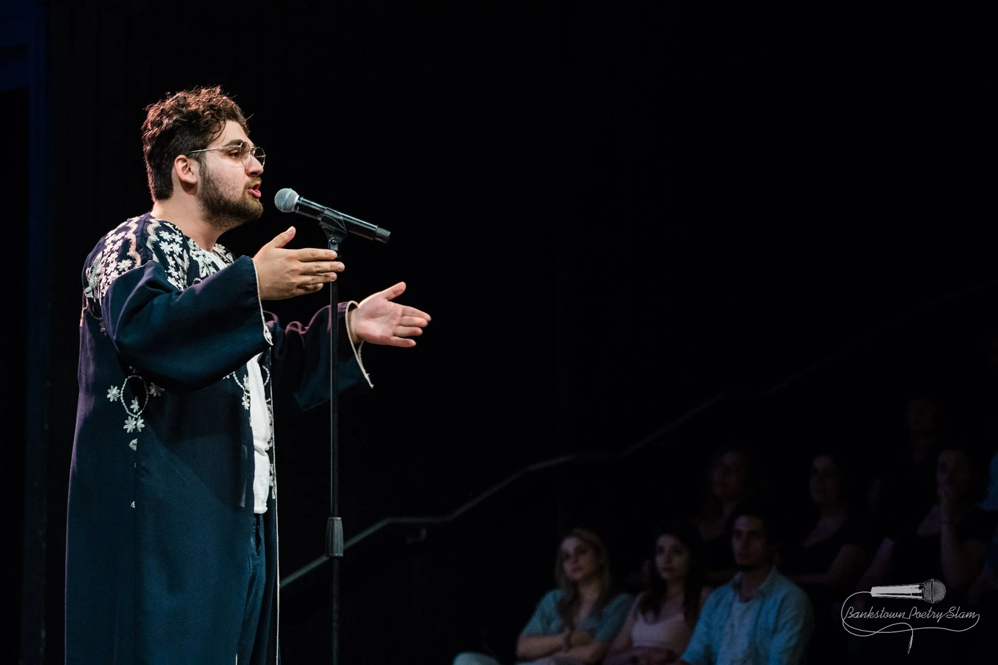 A man with glasses and a beard, wearing a dark floral embroidered shirt, is performing at a poetry slam to an audience.