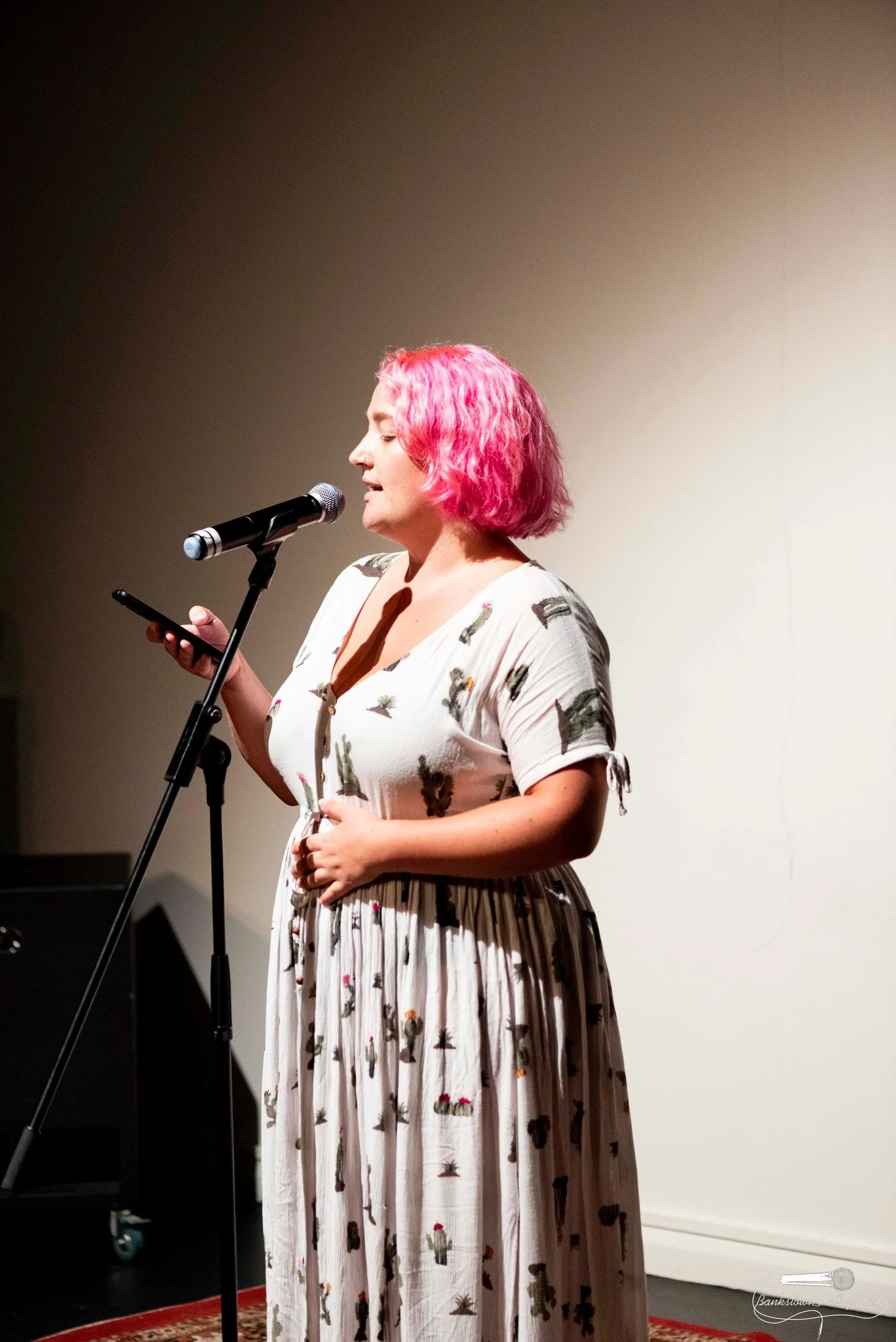 A woman with pink hair standing at a microphone, wearing a white dress with a cactus print, reading from a smartphone against a plain wall.