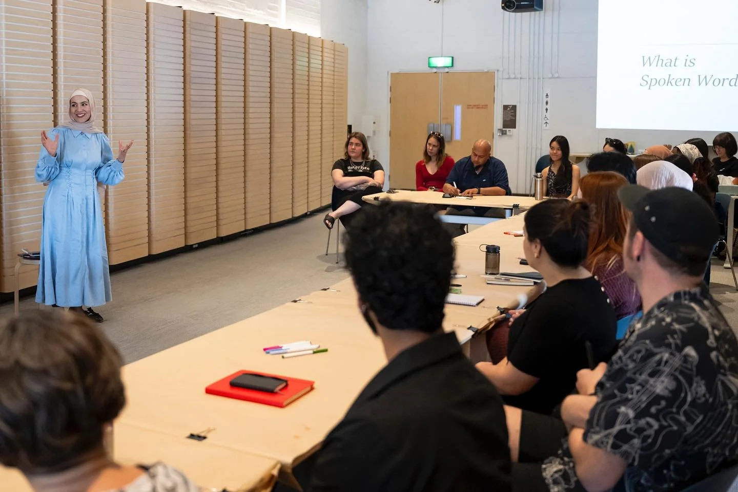 A woman in a long light blue dress and headscarf standing in front of a group of people seated at tables, conducting a poetry workshop.