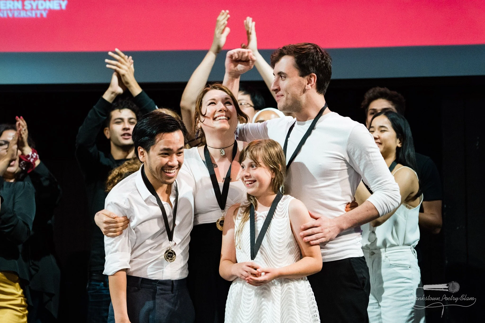 Group of people on stage celebrating, with medals around their necks, some raising their hands, in a large hall or auditorium.