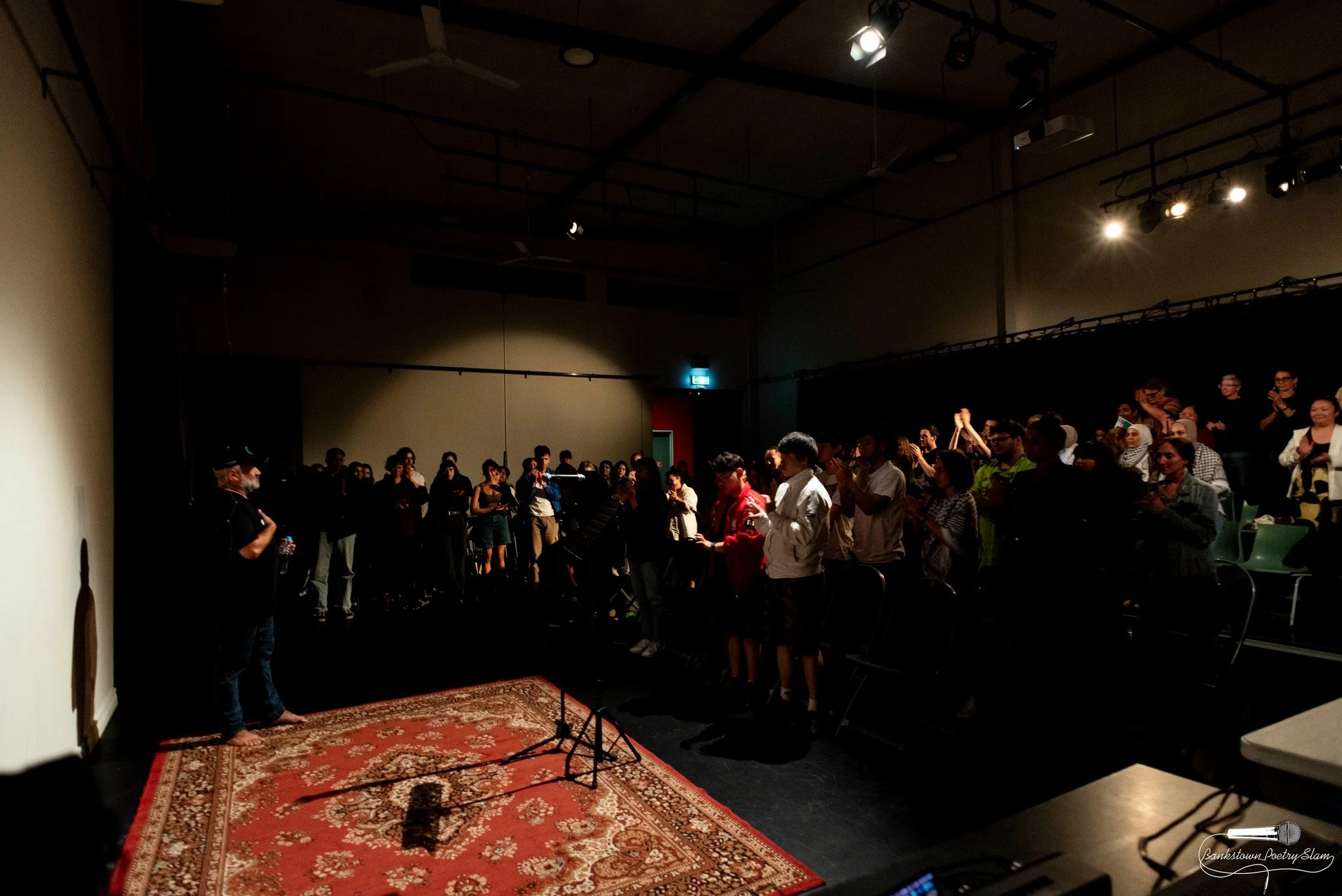 People standing and clapping on a stage and in front of an audience in a dark auditorium. There is a red patterned rug on the stage, microphones, and stage lighting.